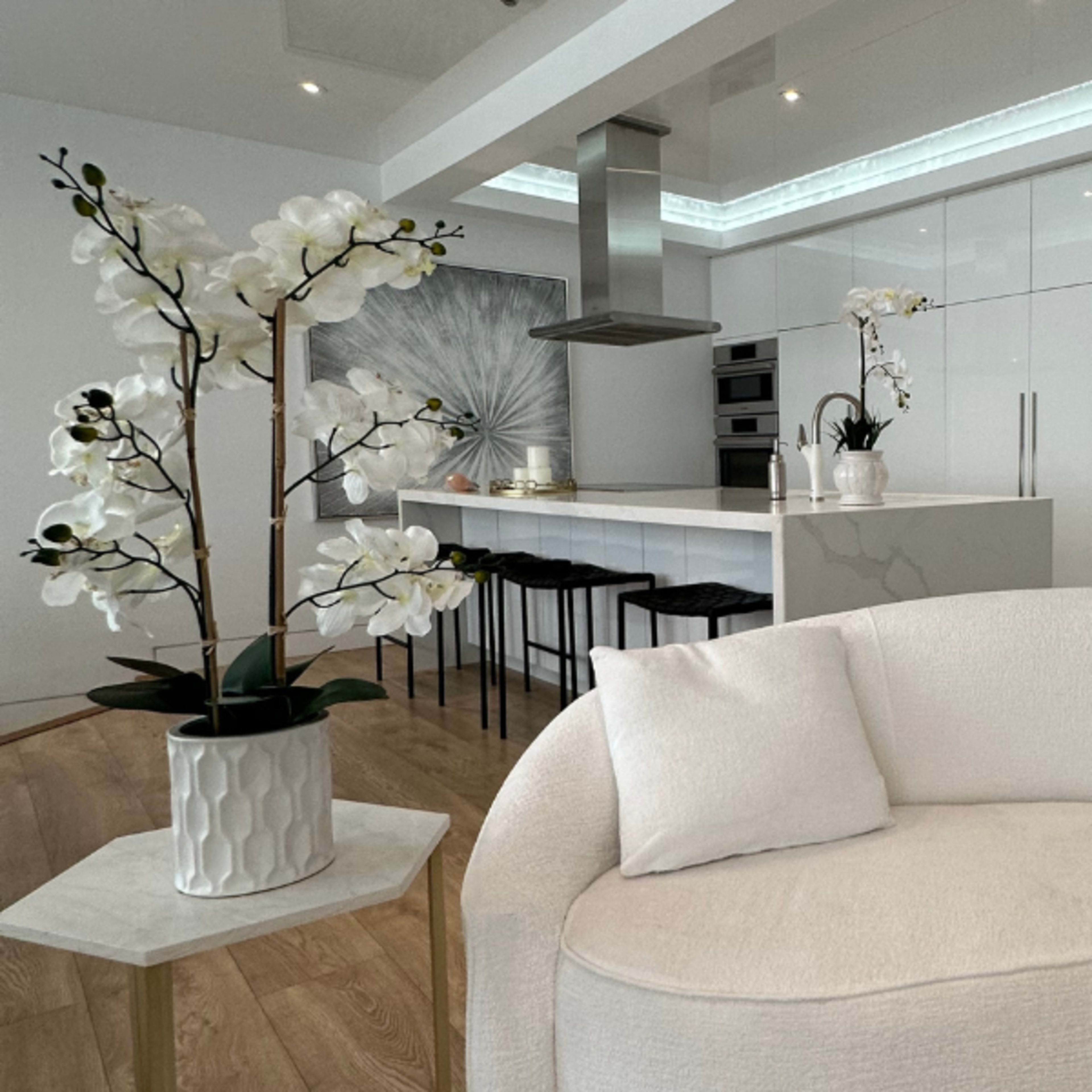 A modern kitchen space features white cabinetry, a central island with bar stools, and decorative orchids in a minimalist living area.