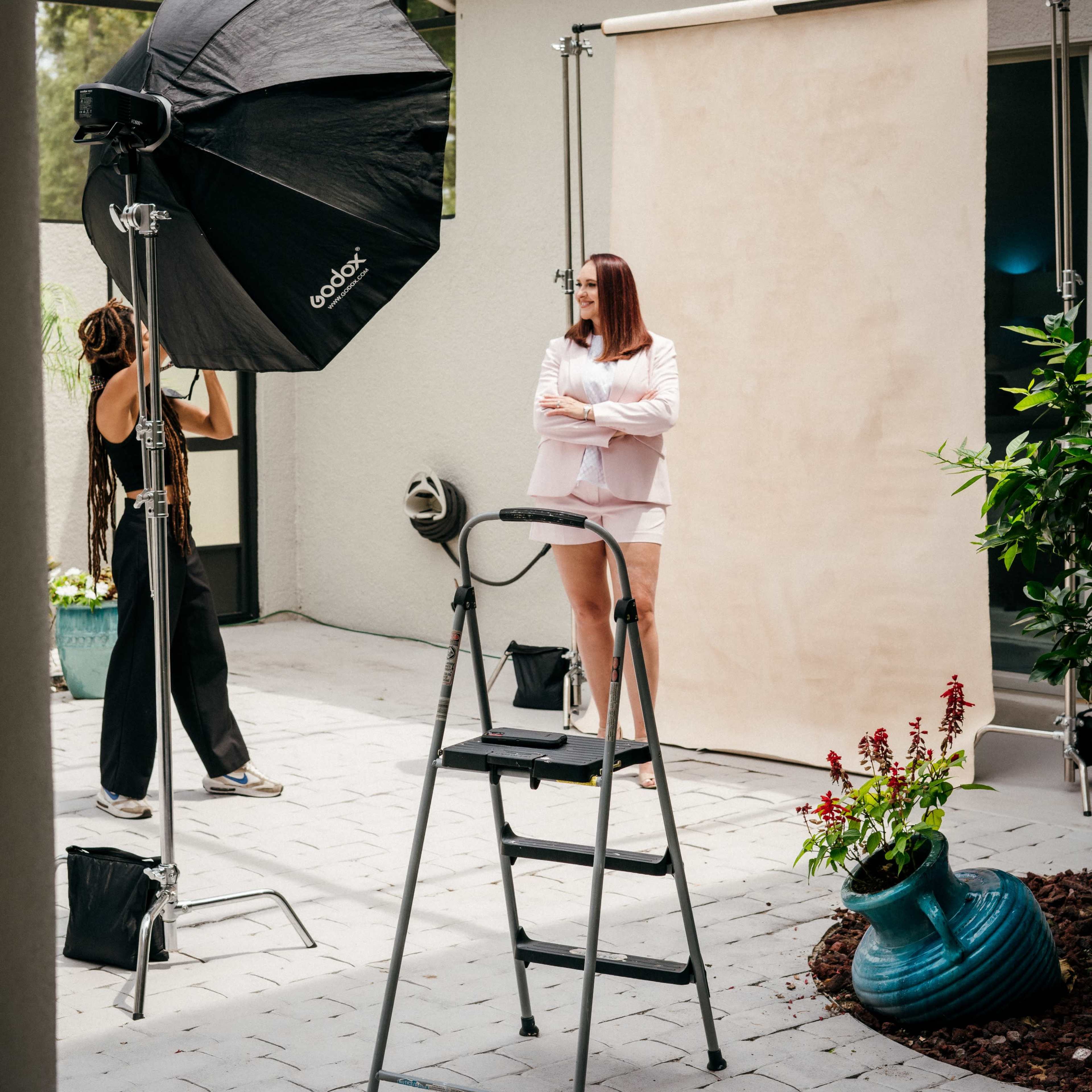 A woman stands on a ladder in front of a backdrop while another person prepares a light setup for a photo shoot in an outdoor setting.