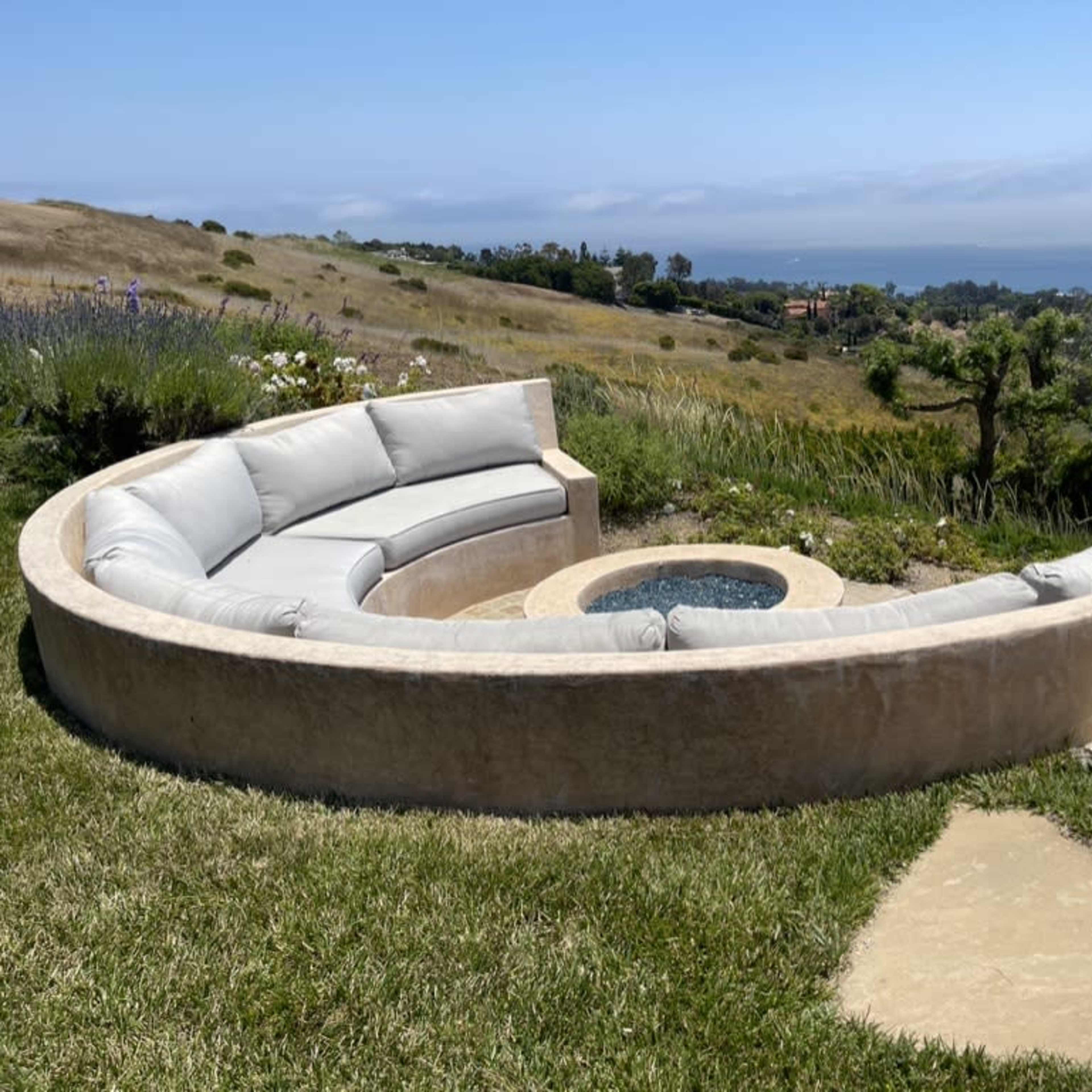 A semicircular outdoor seating area surrounds a small fire pit, overlooking a grassy hillside and distant ocean view.