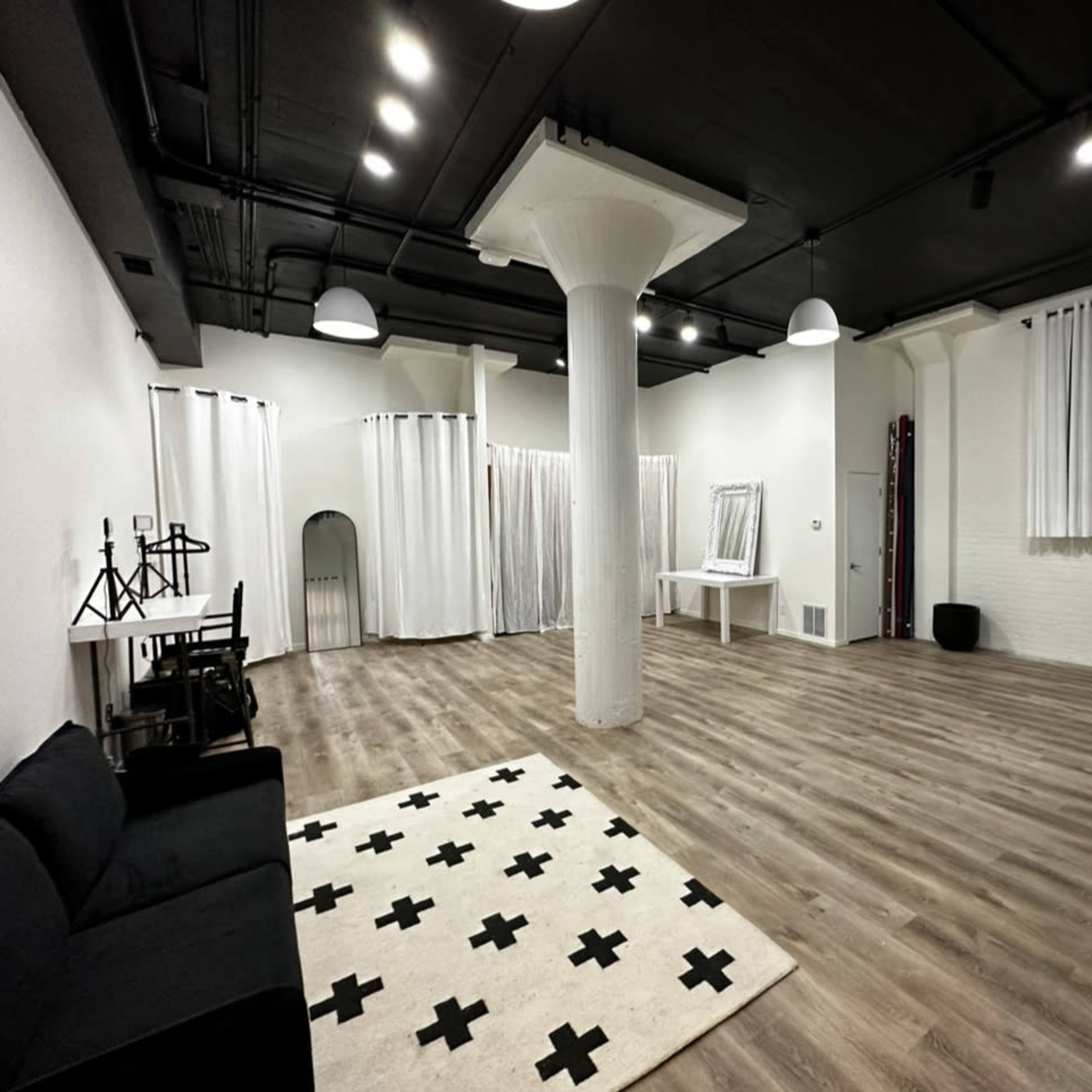 A spacious, empty room with wooden flooring, a white wall, a sofa, and a large area rug featuring a black cross pattern.