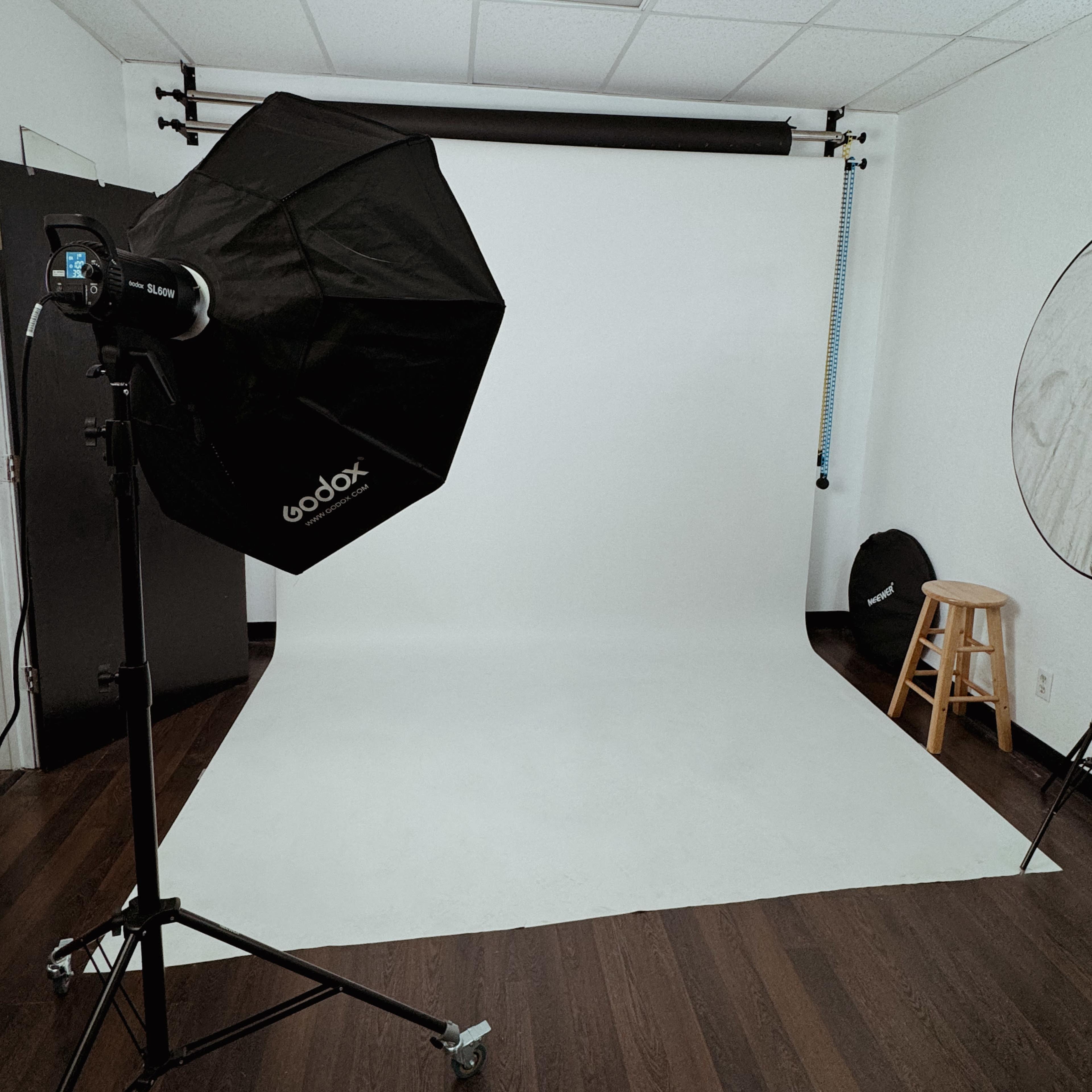A photography studio setup with a softbox light, a white backdrop, and a stool against a wooden floor.