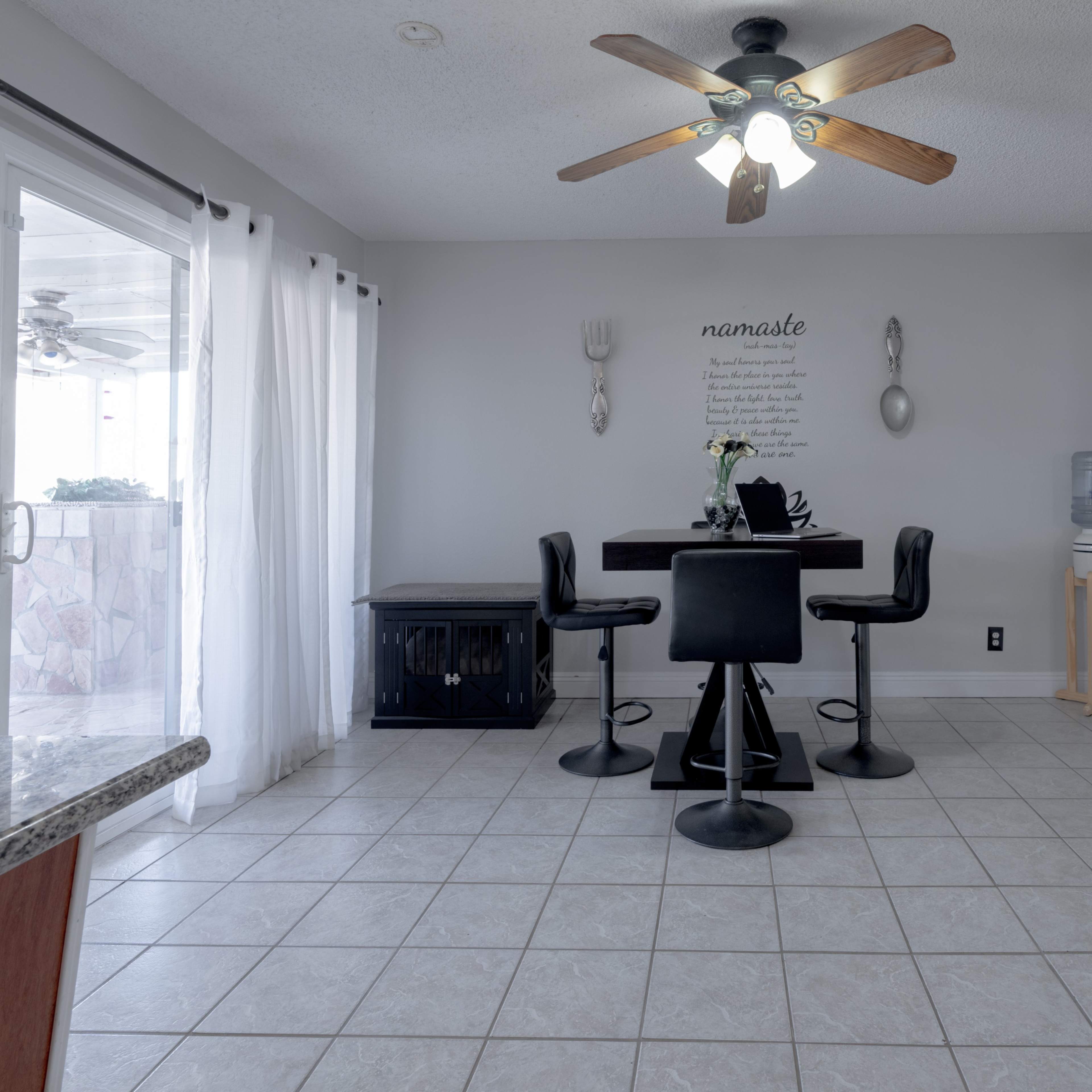 A dining area features a round black table with four chairs, a ceiling fan, and light-colored walls with decorative wall art.