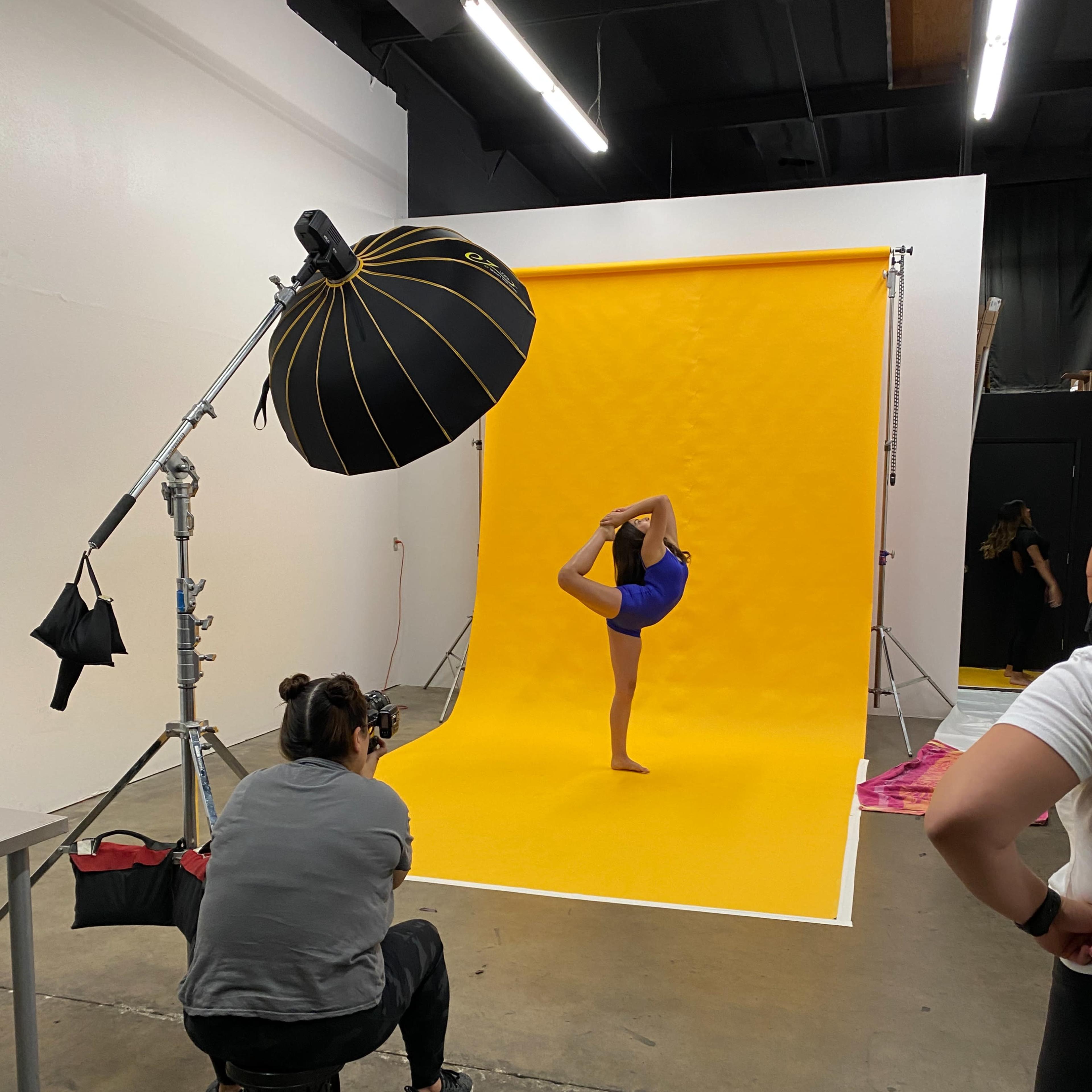 A dancer performs a pose on one leg in front of a yellow backdrop as a photographer captures the image.
