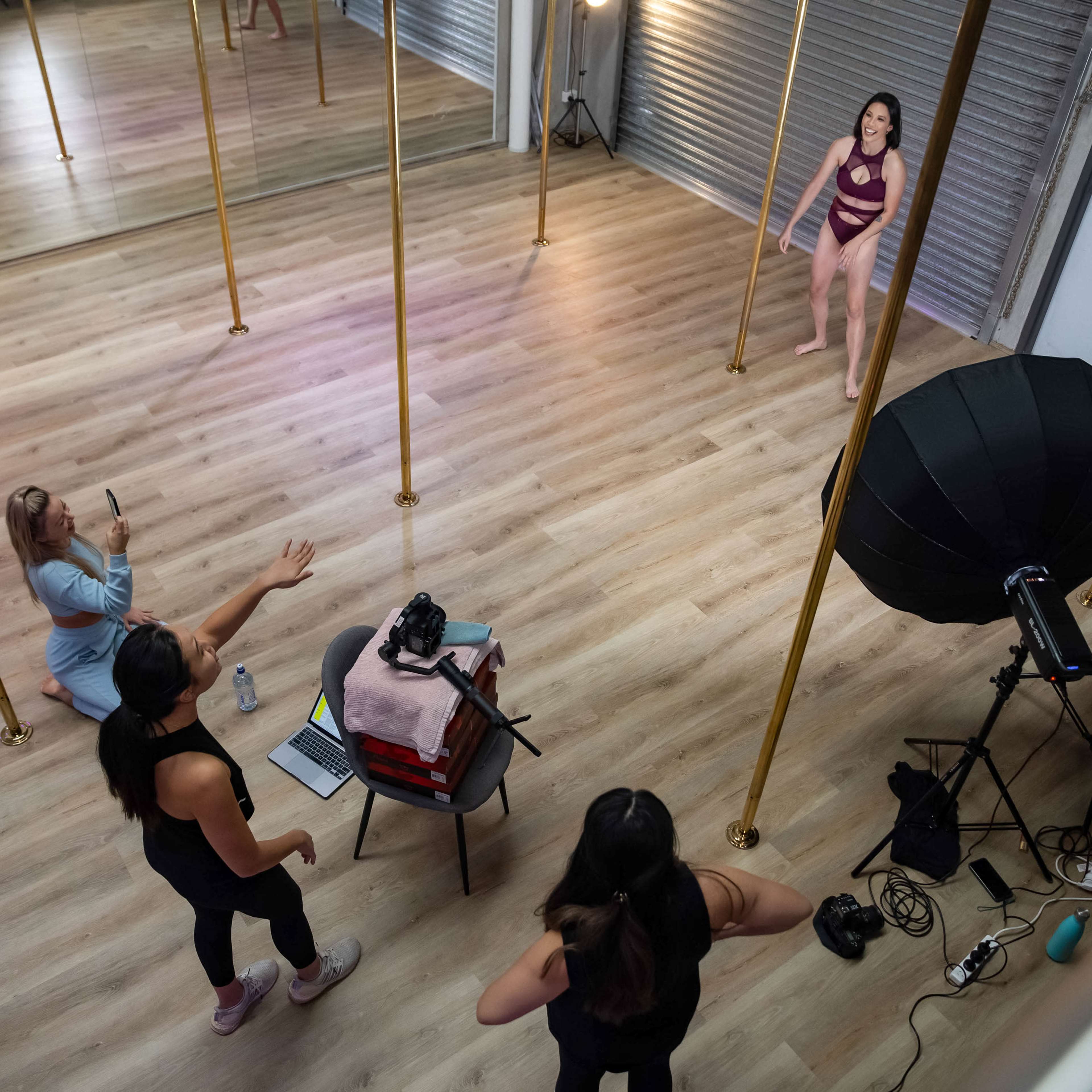 A studio with four women, two standing near a pole while one is sitting behind a camera, and another is gesturing from the side.