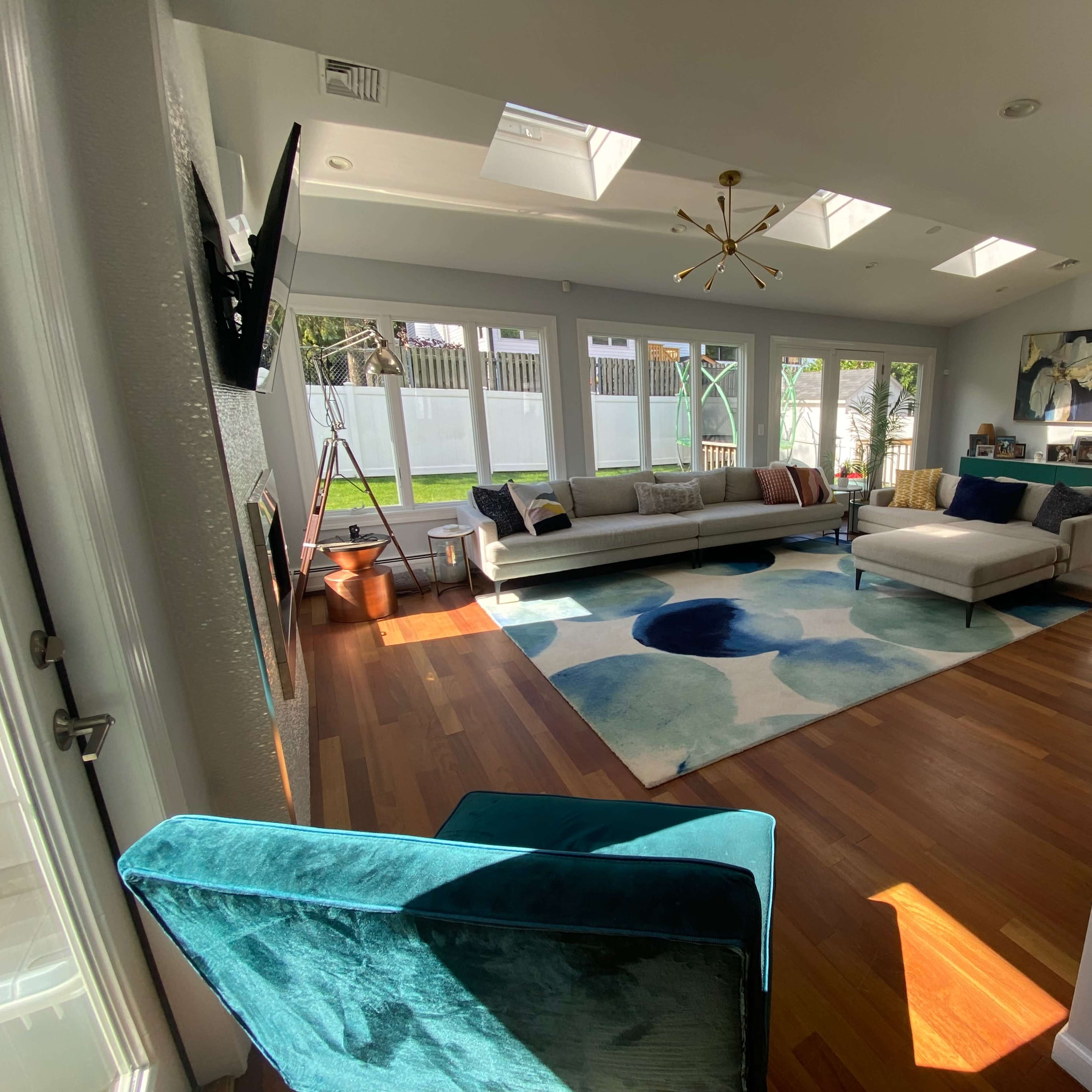 A spacious living room with large windows, a soft area rug featuring a colorful design, and a sectional sofa arranged around a central coffee table.