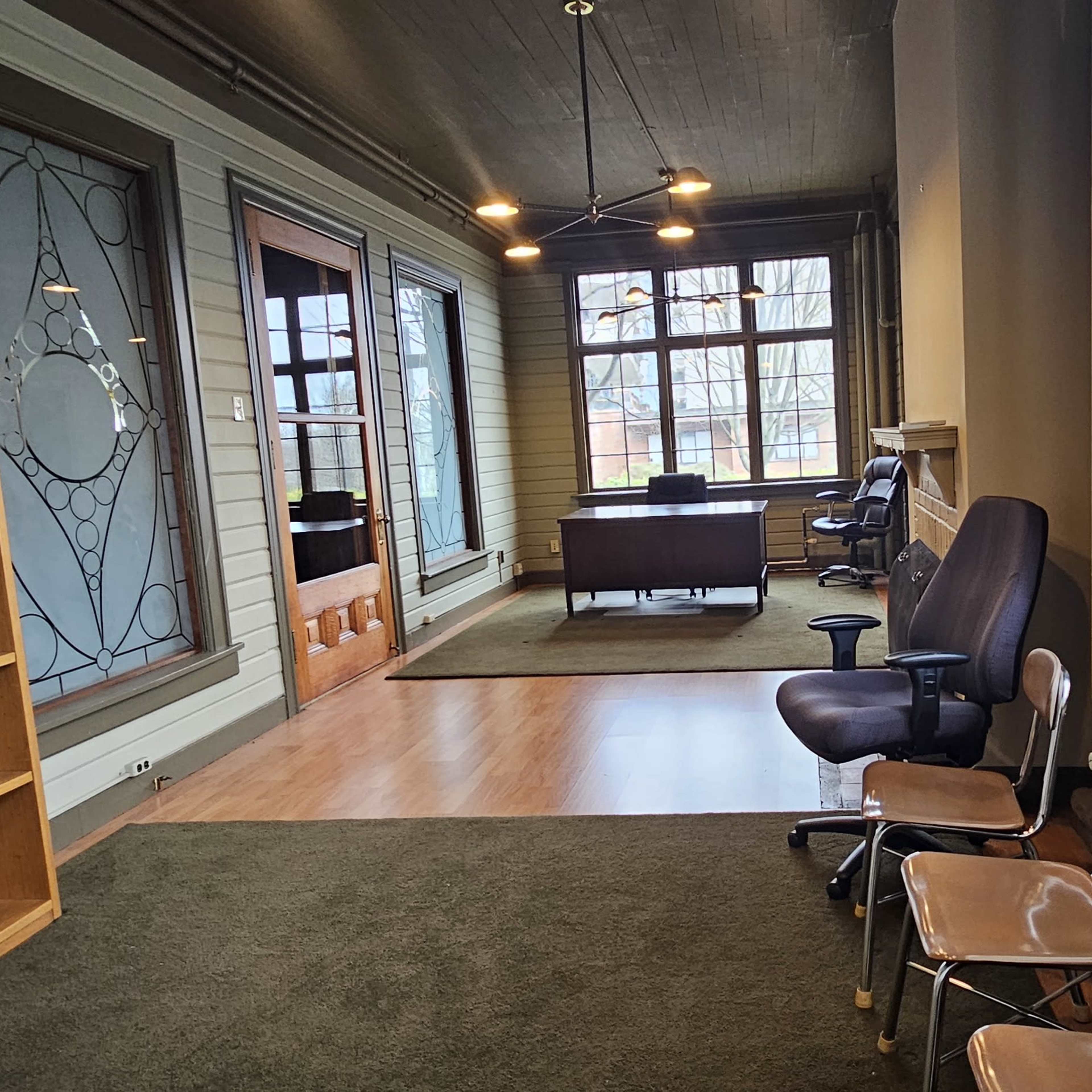 A hallway with a wooden bookshelf, several chairs, and large windows illuminating a desk area in the background.