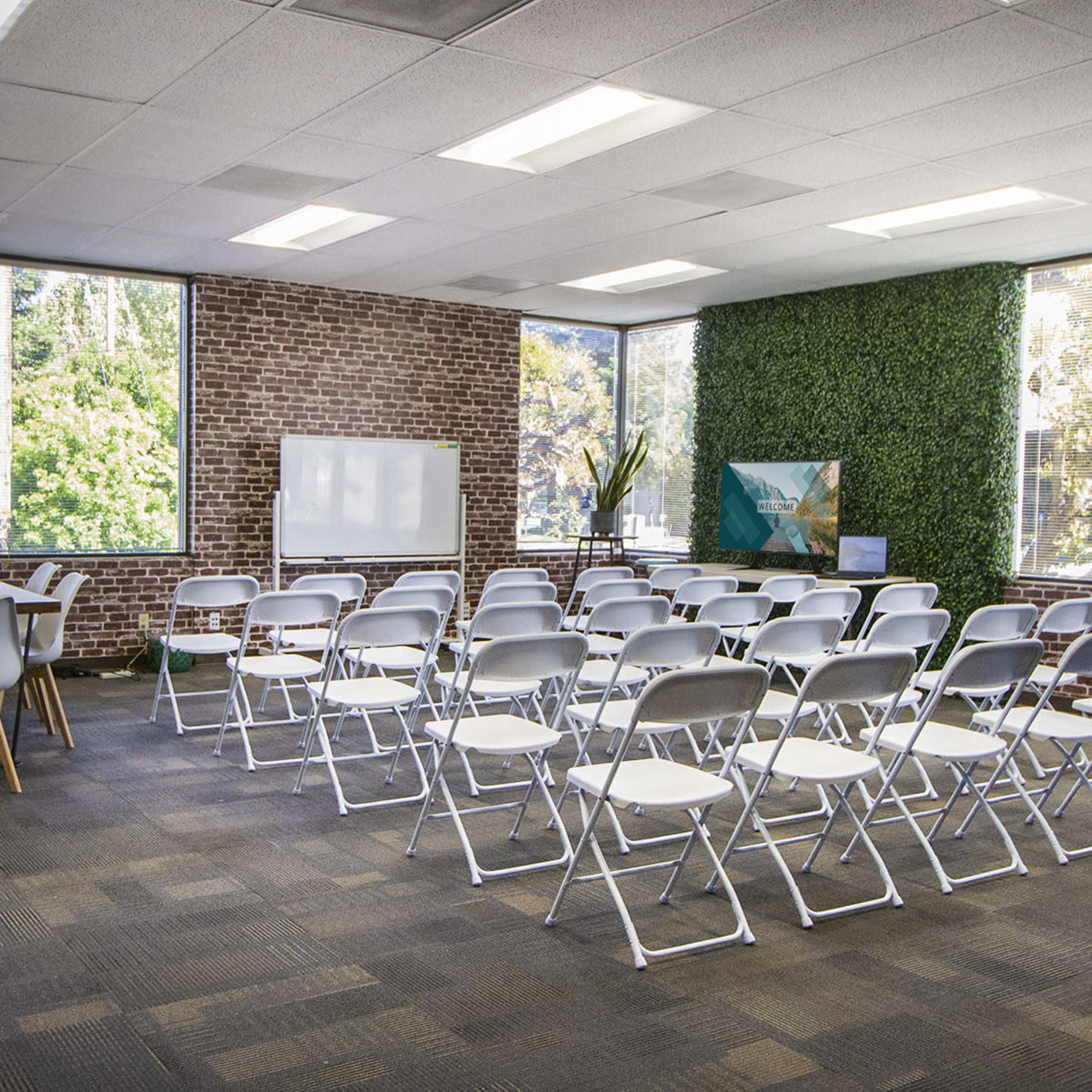 A well-lit meeting room with rows of white folding chairs arranged facing a whiteboard, surrounded by large windows and greenery on the walls.
