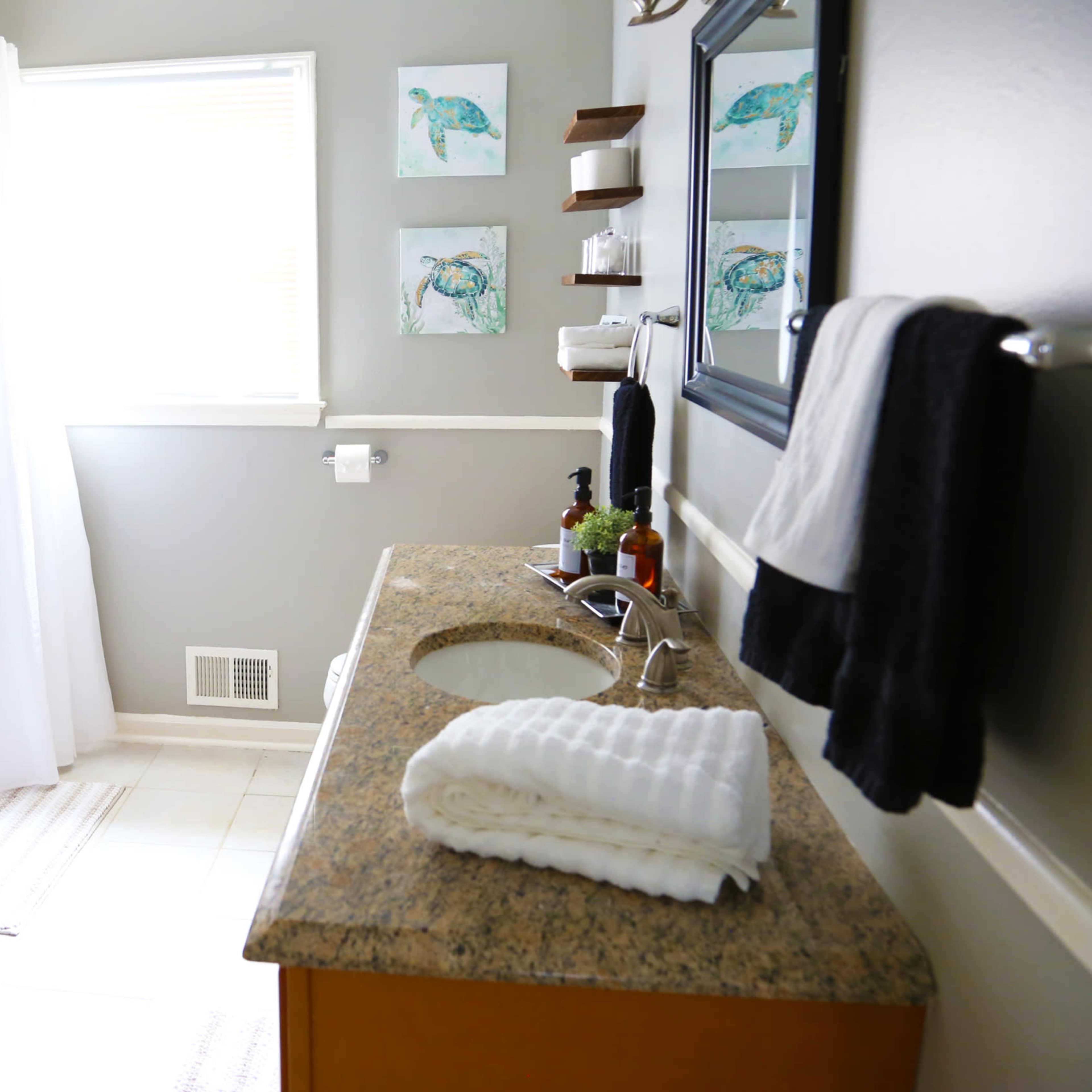 A clean bathroom features a tub with a shower curtain, a granite countertop with neatly stacked towels, and fish-themed artwork on the wall.