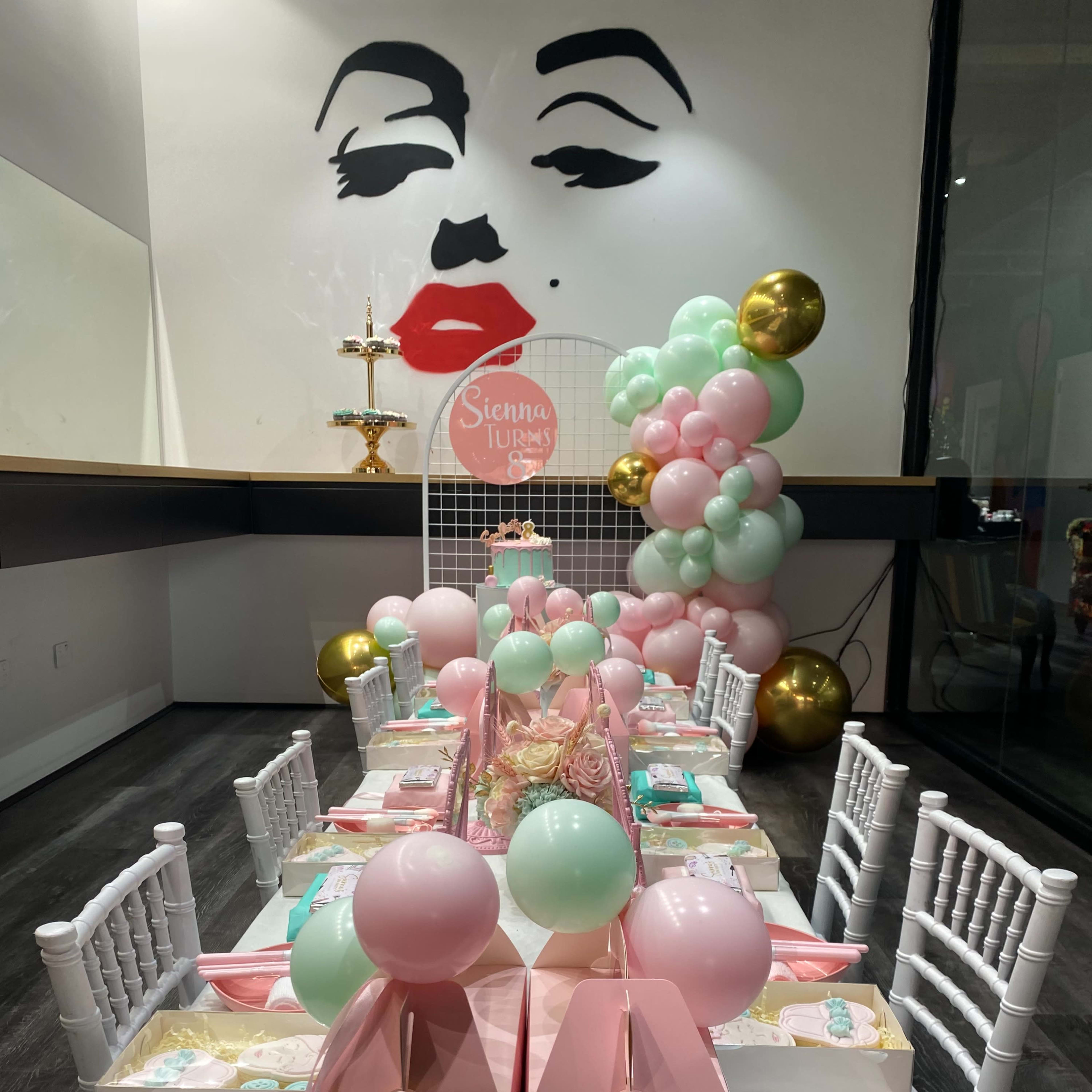 A long table is set for a party, adorned with pastel-colored decorations, gift boxes, and balloons, against a decorative wall featuring a large mural of a woman's face.