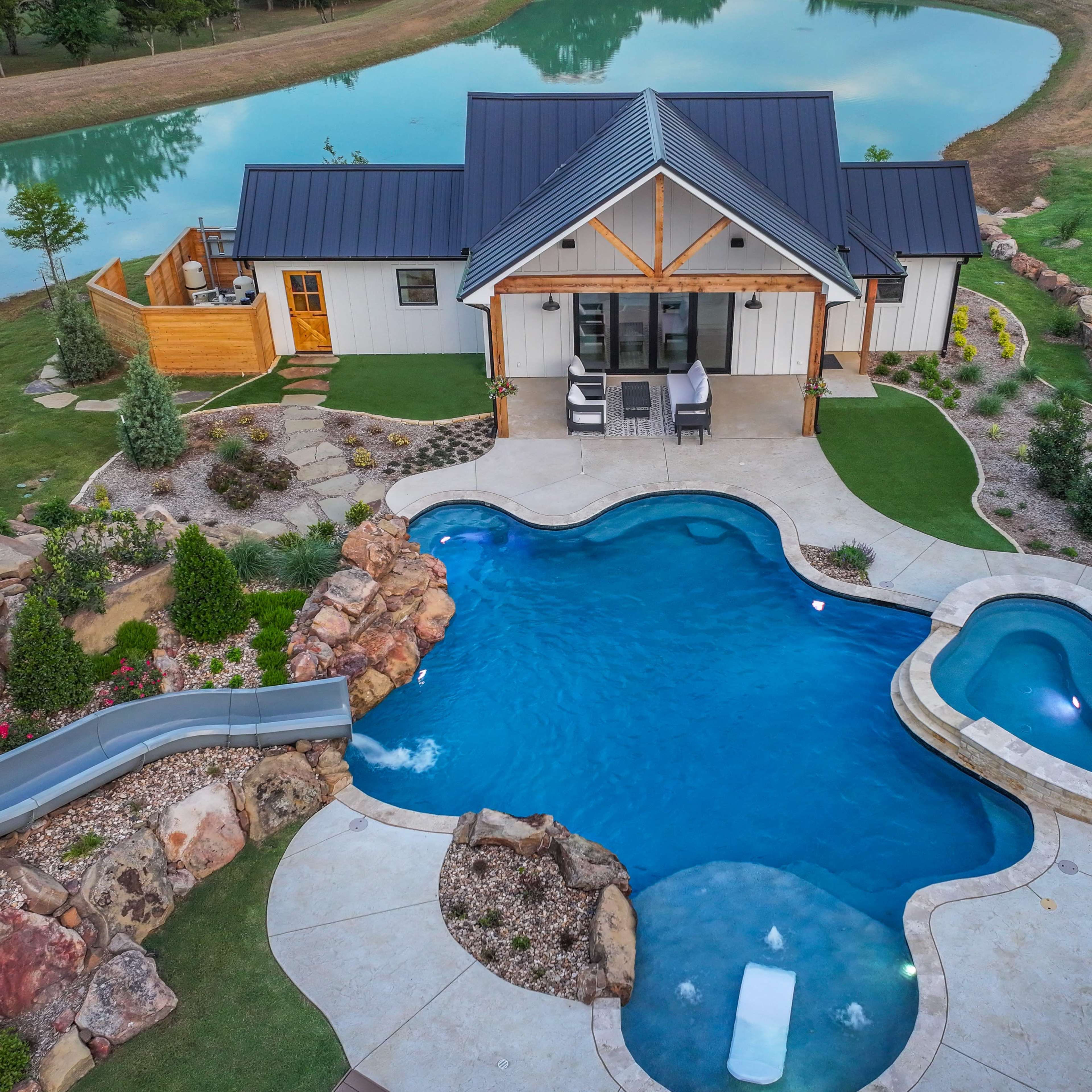 Aerial view of a house situated by a pond, featuring a curvy swimming pool and a separate spa area surrounded by landscaping.