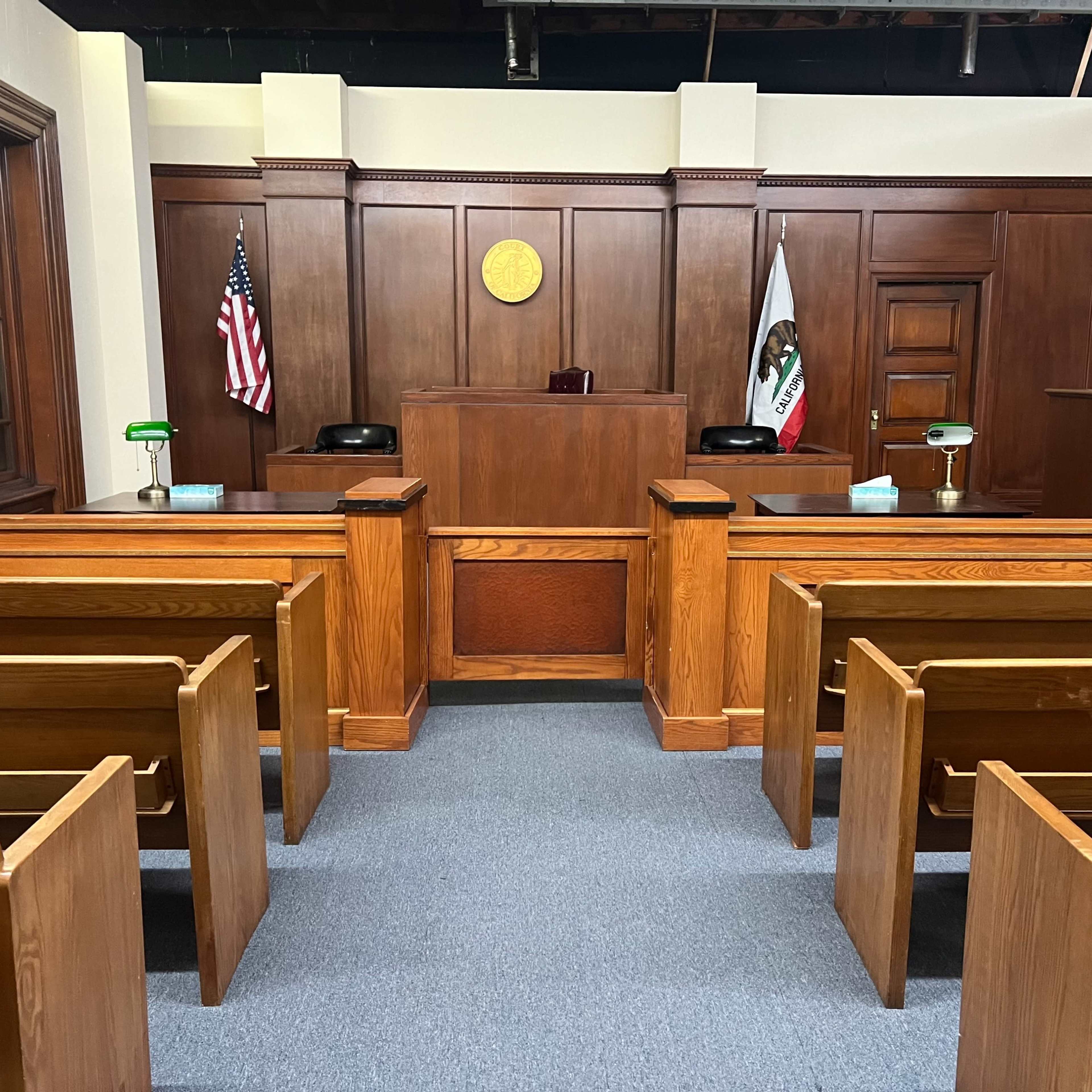 A courtroom interior with wooden benches, a judge's bench at the front, and flags positioned by the side walls.