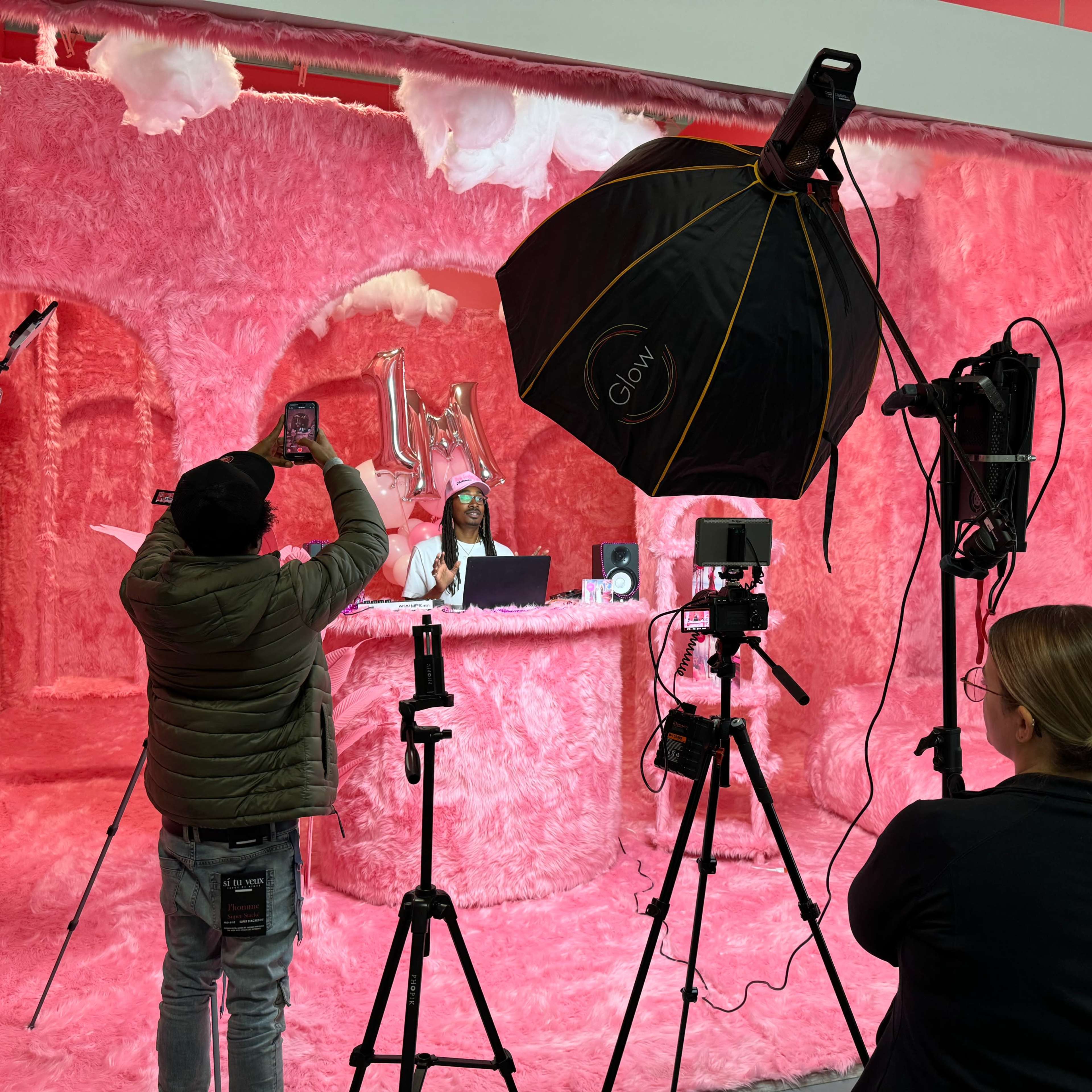 A photoshoot is taking place in a pink, furry studio with a person behind a desk and two individuals capturing images with cameras.