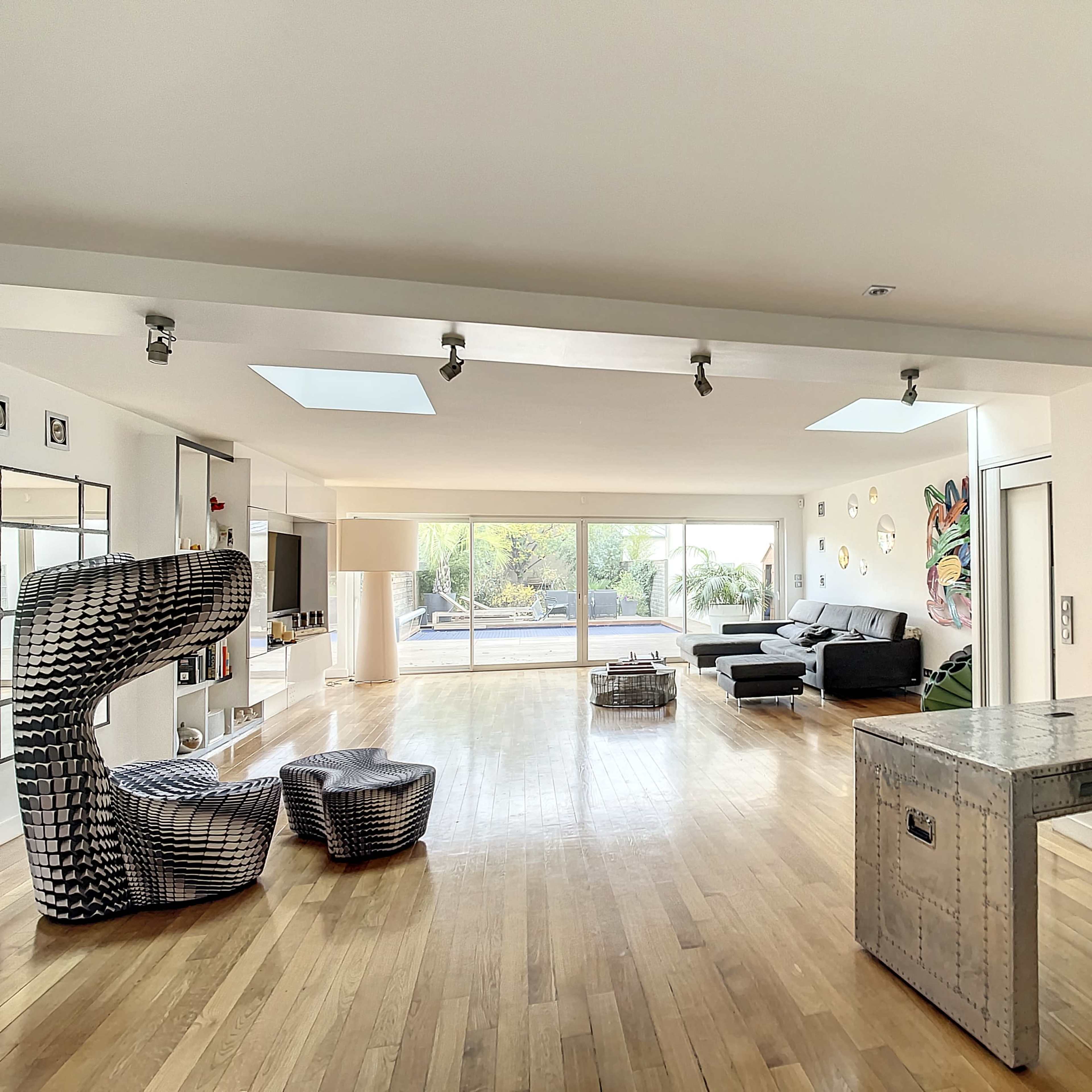 A spacious, modern living area with wooden flooring, a large window allowing natural light, and contemporary furniture pieces, including a unique chair and a metallic table.