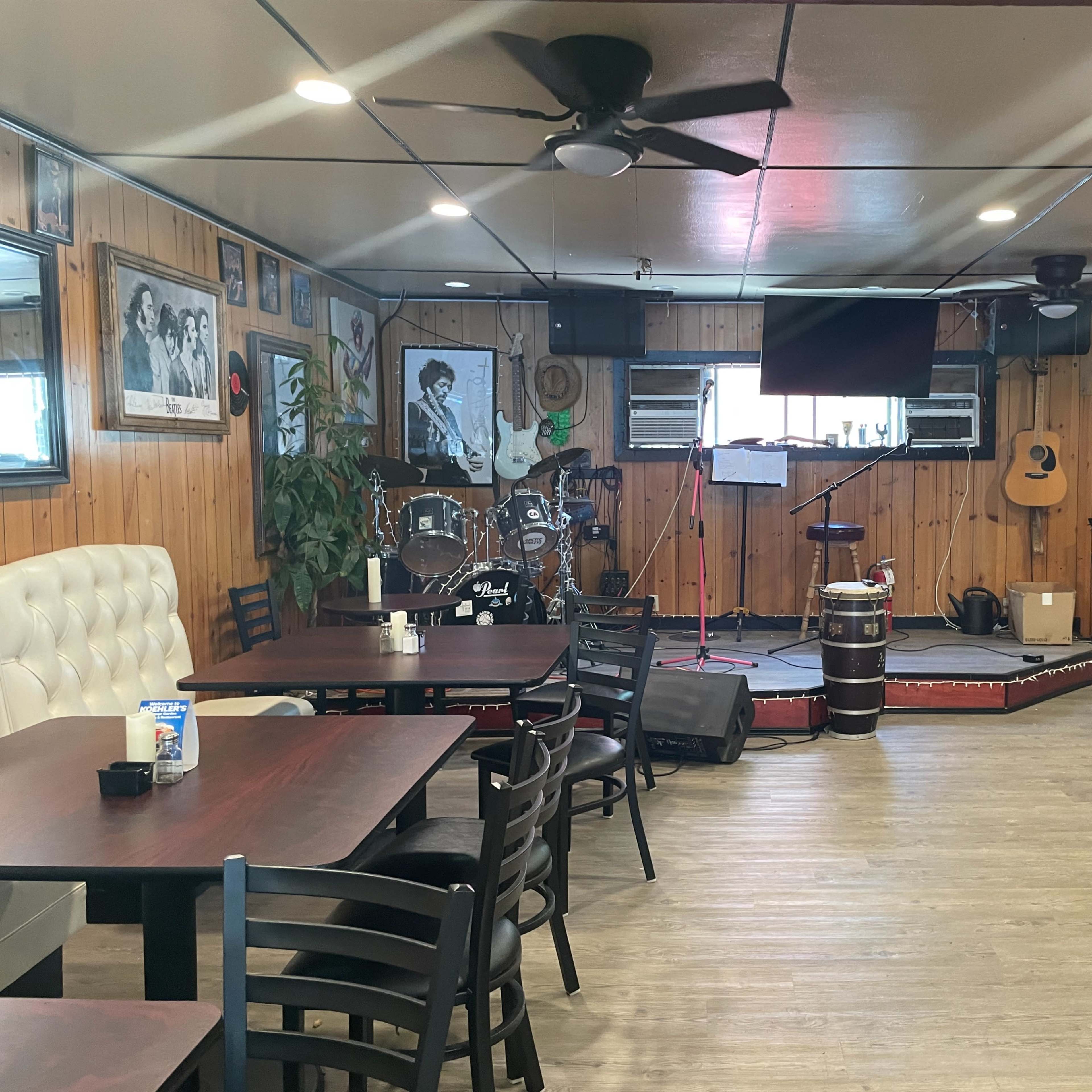 An interior of a restaurant or bar with wooden walls, tables arranged for dining, and a small stage equipped with musical instruments.