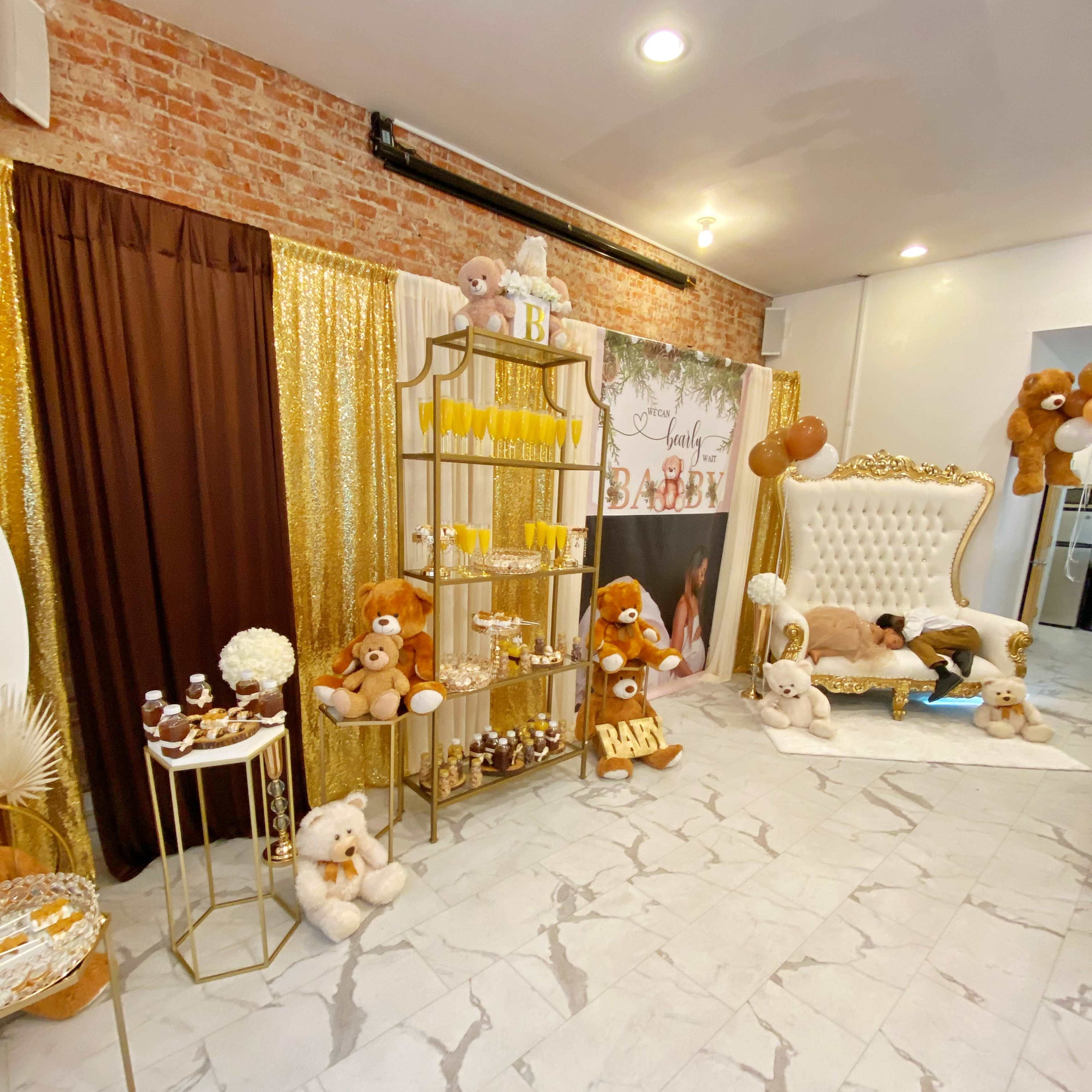 A decorated event space featuring a gold and brown color scheme, with plush teddy bears, a dessert table, and a large white chair against a brick wall backdrop.