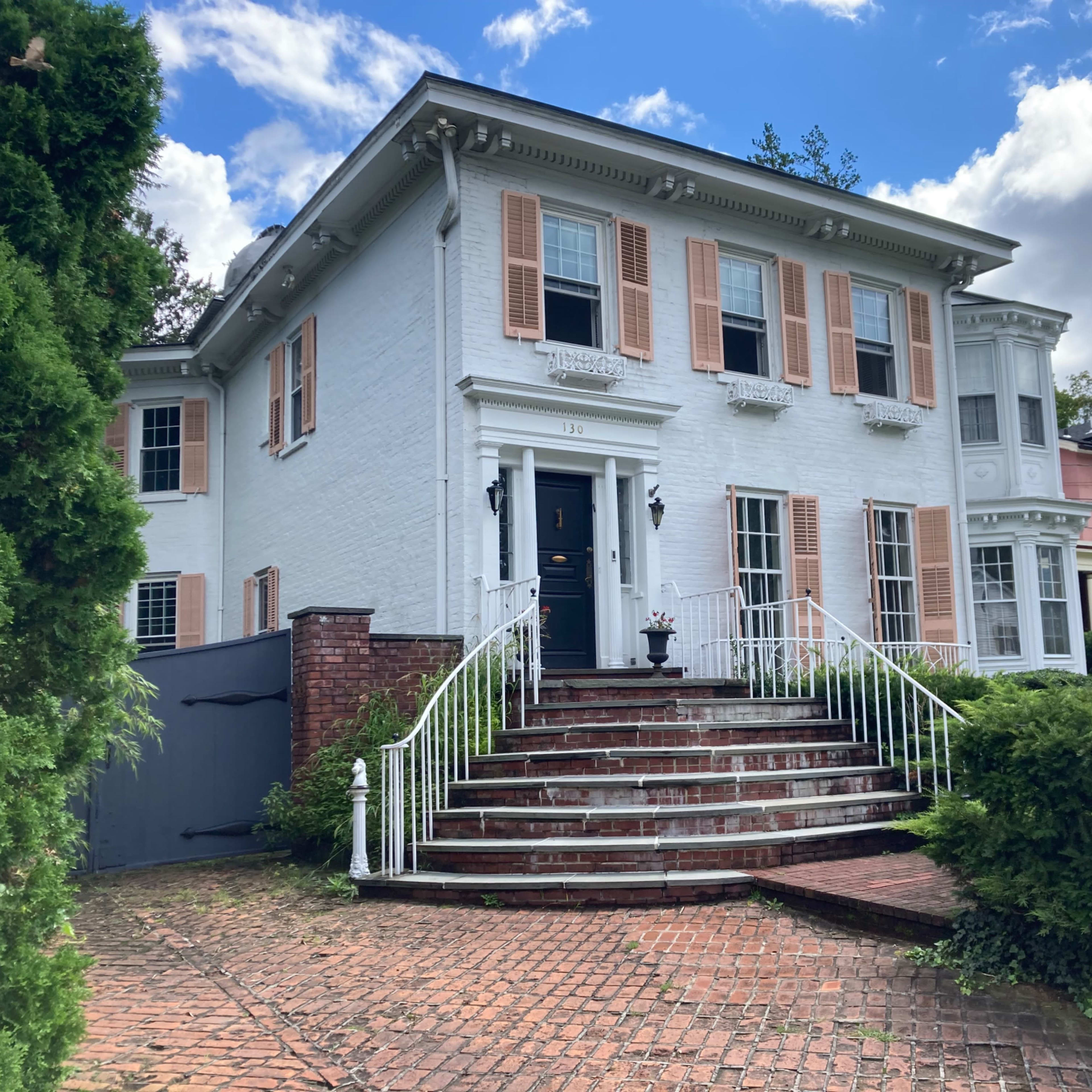 A large white house with brick steps, a black front door, and decorative shutters, flanked by green shrubs and surrounded by other residential buildings.
