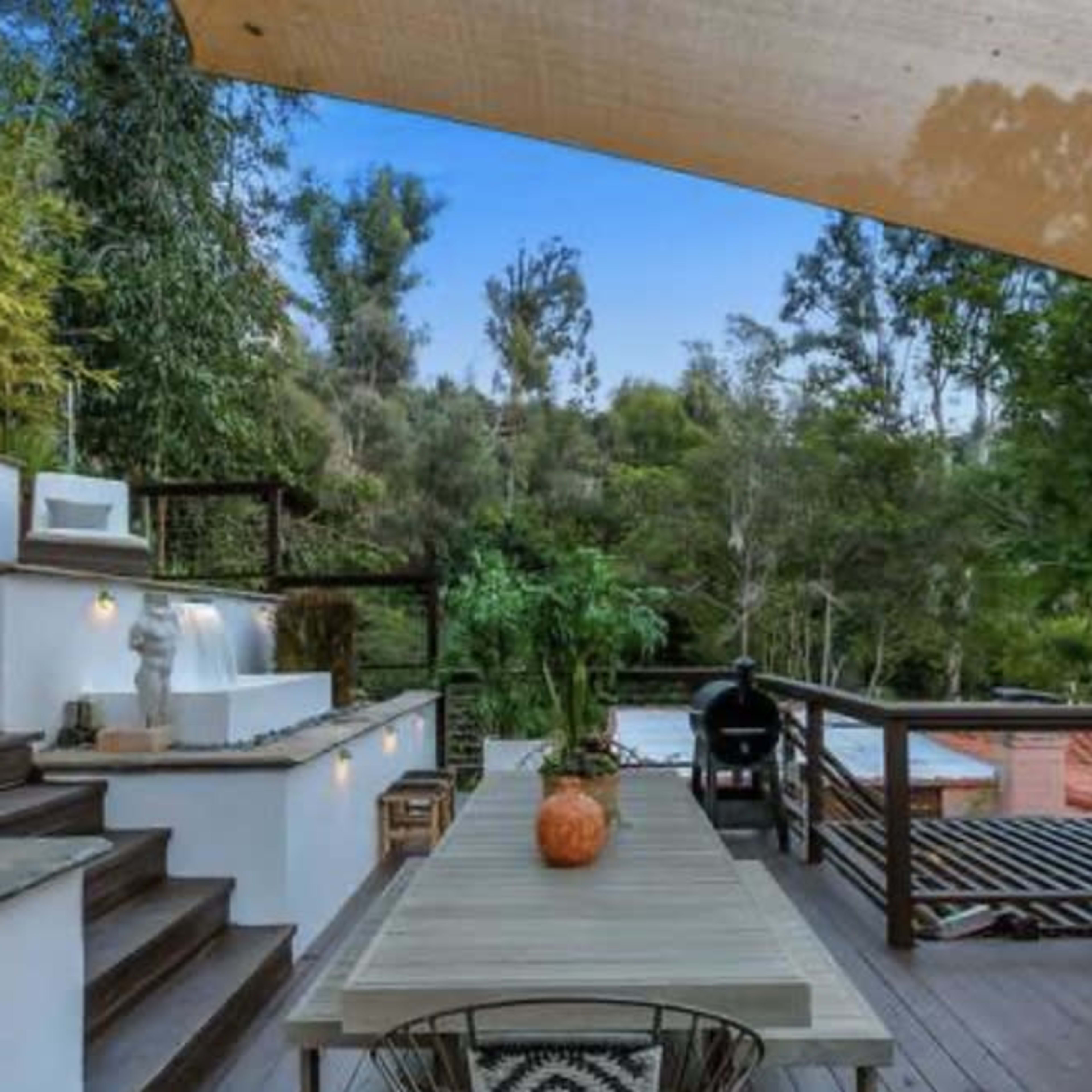A modern outdoor patio features a wooden table surrounded by greenery, with steps leading to a second level and a distant pool area.