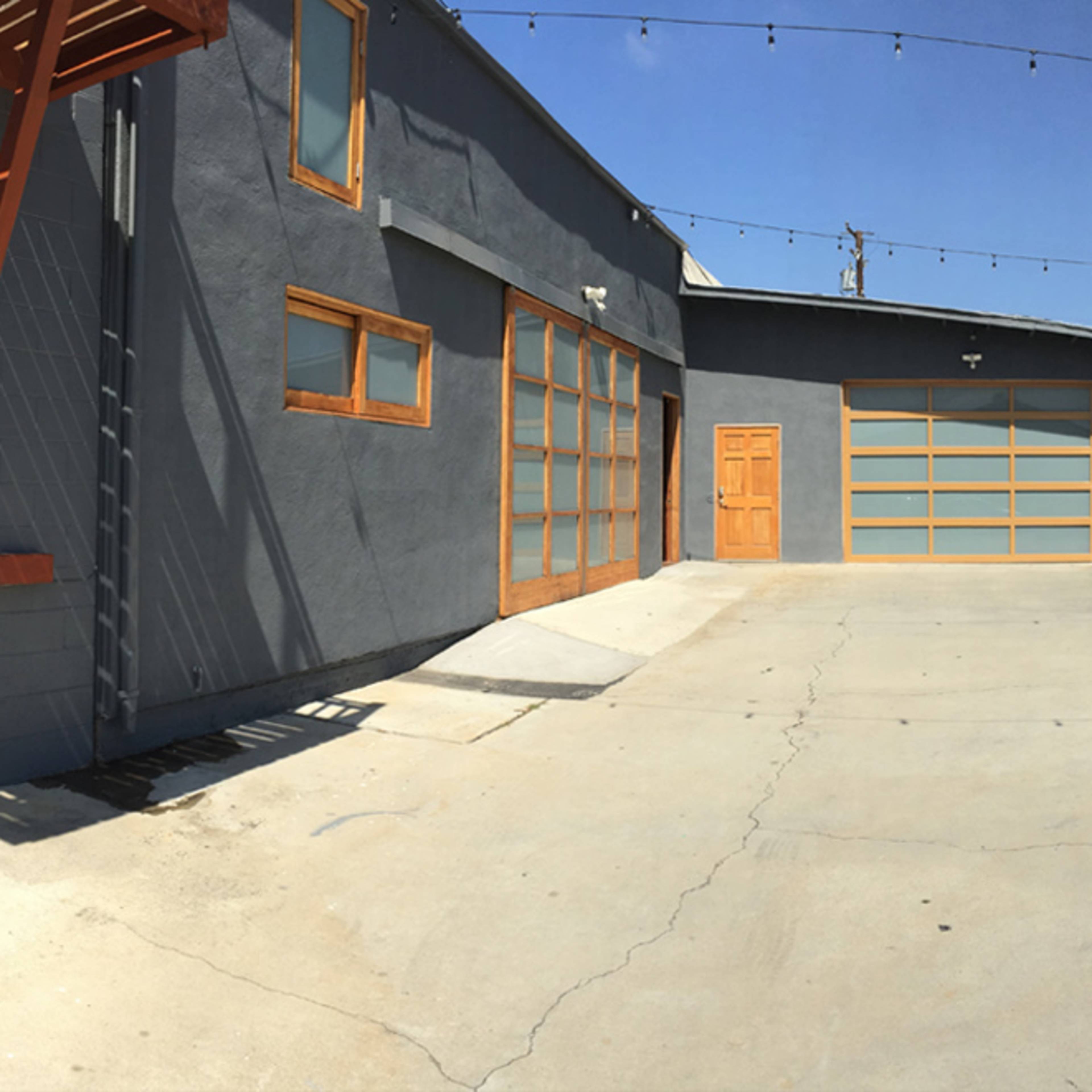 A panoramic view of a gray building with wooden accents and a large paved parking area, surrounded by low wooden fences and string lights overhead.