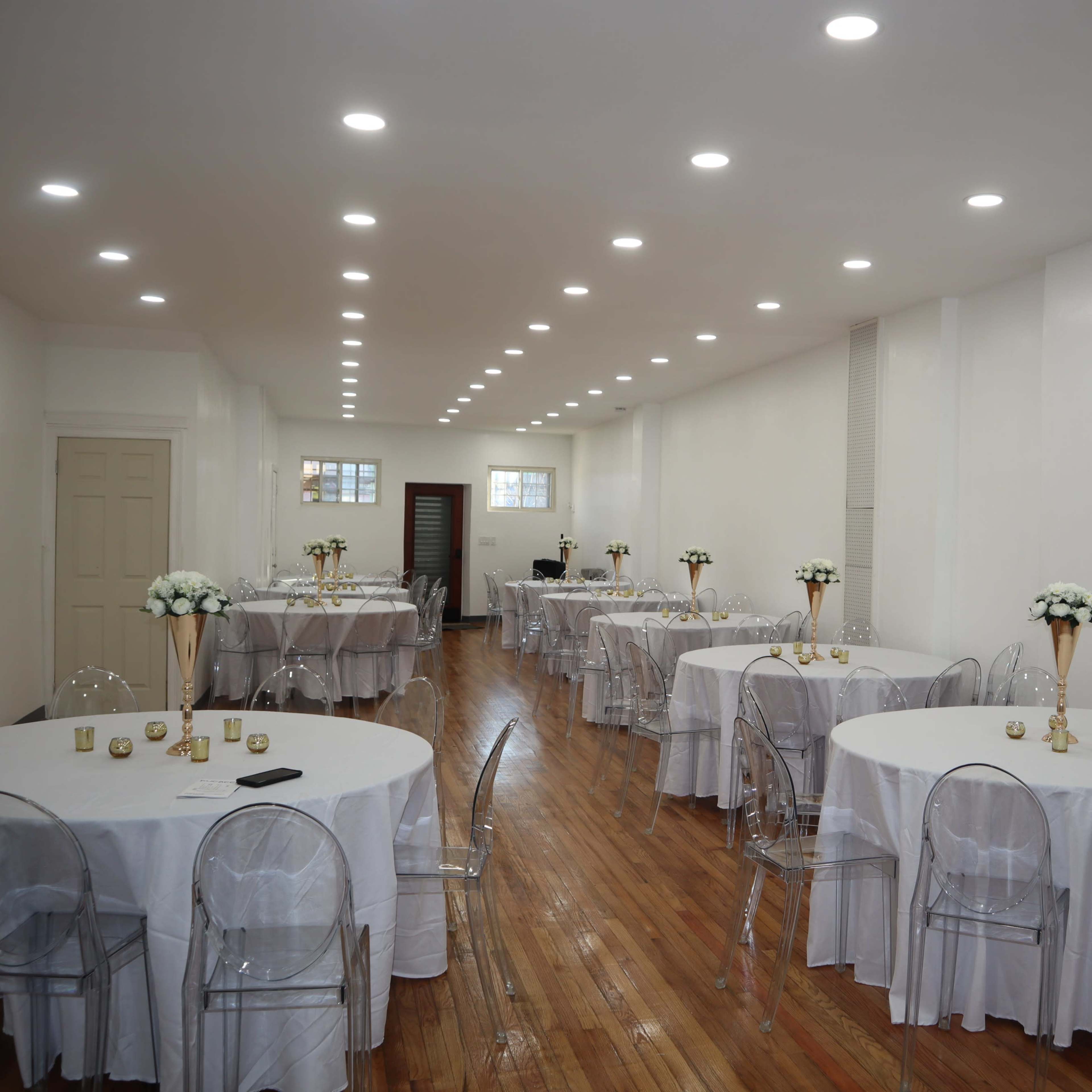 A well-lit banquet hall features several round tables with white tablecloths and clear plastic chairs arranged along a polished wooden floor.