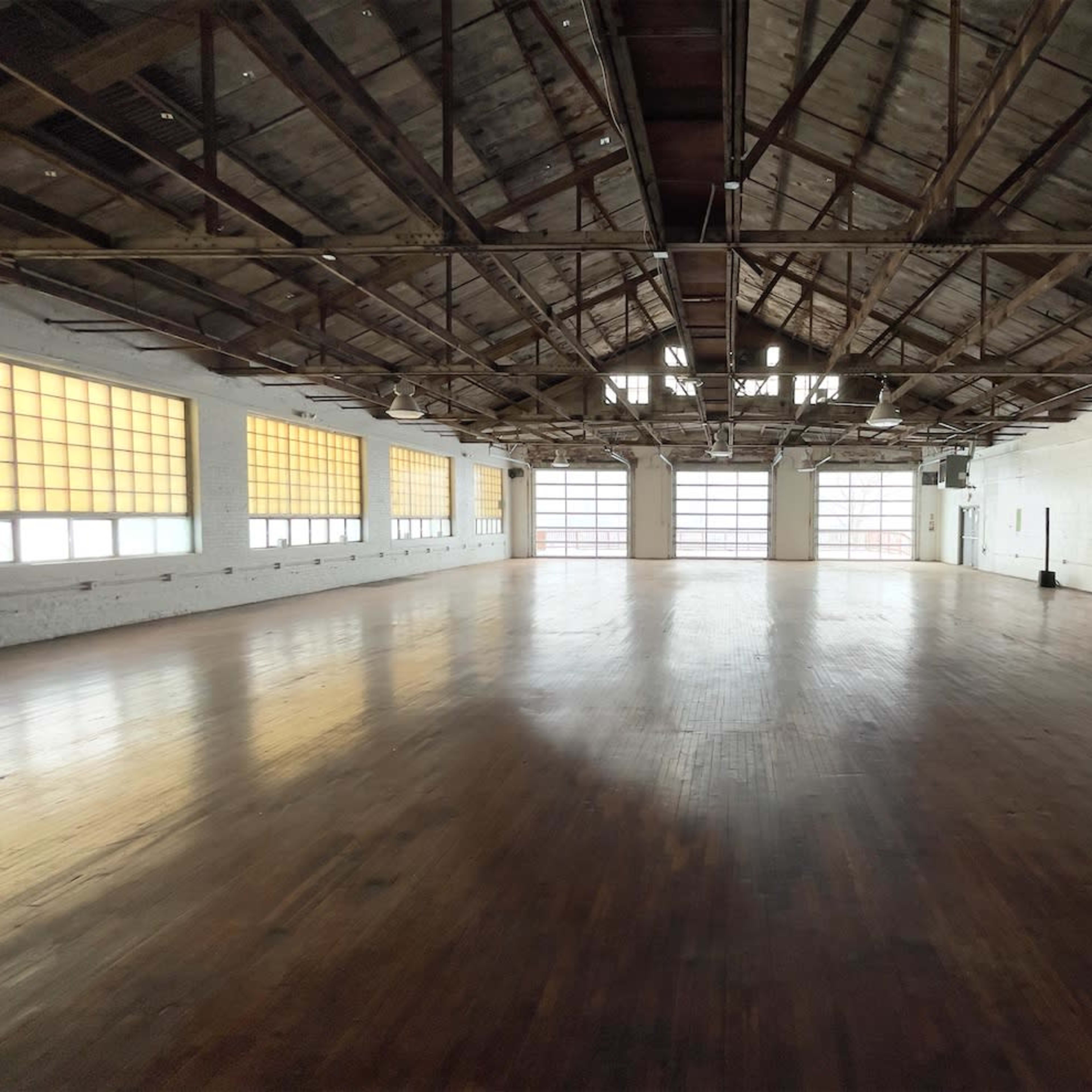 A spacious, empty room with wooden floors and industrial-style windows along the walls, featuring exposed beams and a high ceiling.