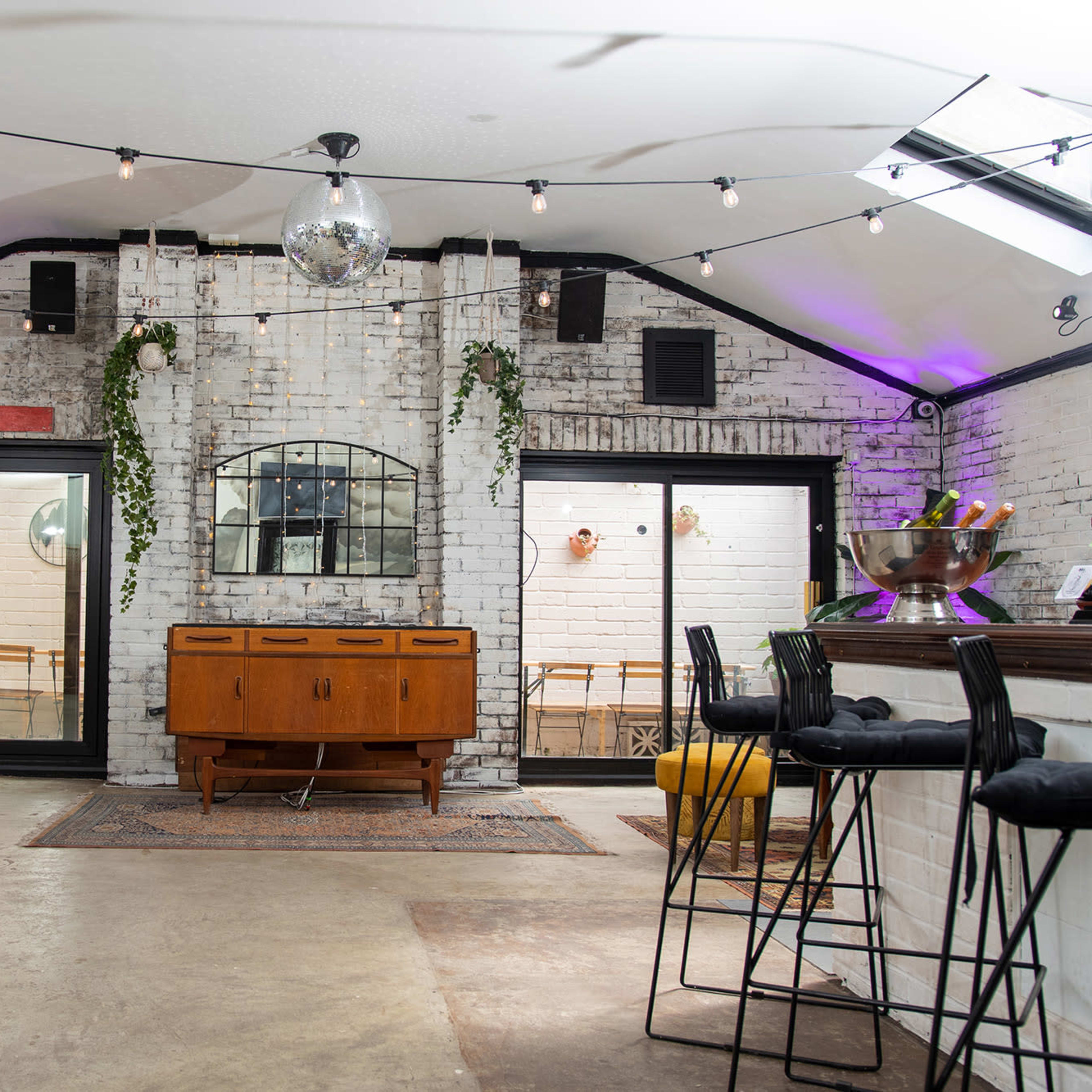 A spacious, modern interior with a bar area, large windows, and decorative lighting, featuring a mix of white brick walls and greenery.