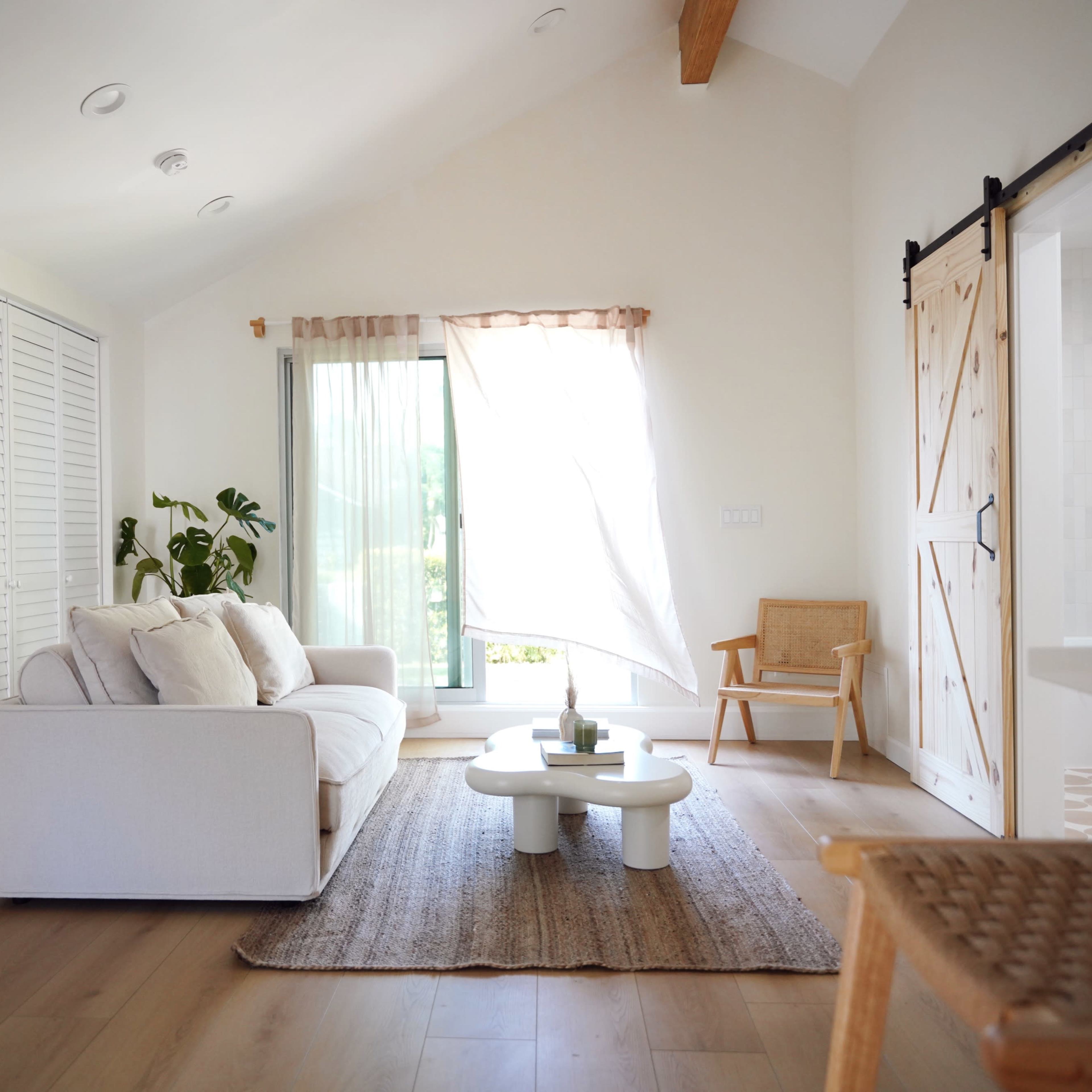 A bright, airy living room features a white sofa, a round coffee table, and large windows with sheer curtains, complemented by a woven rug and a wooden sliding door.