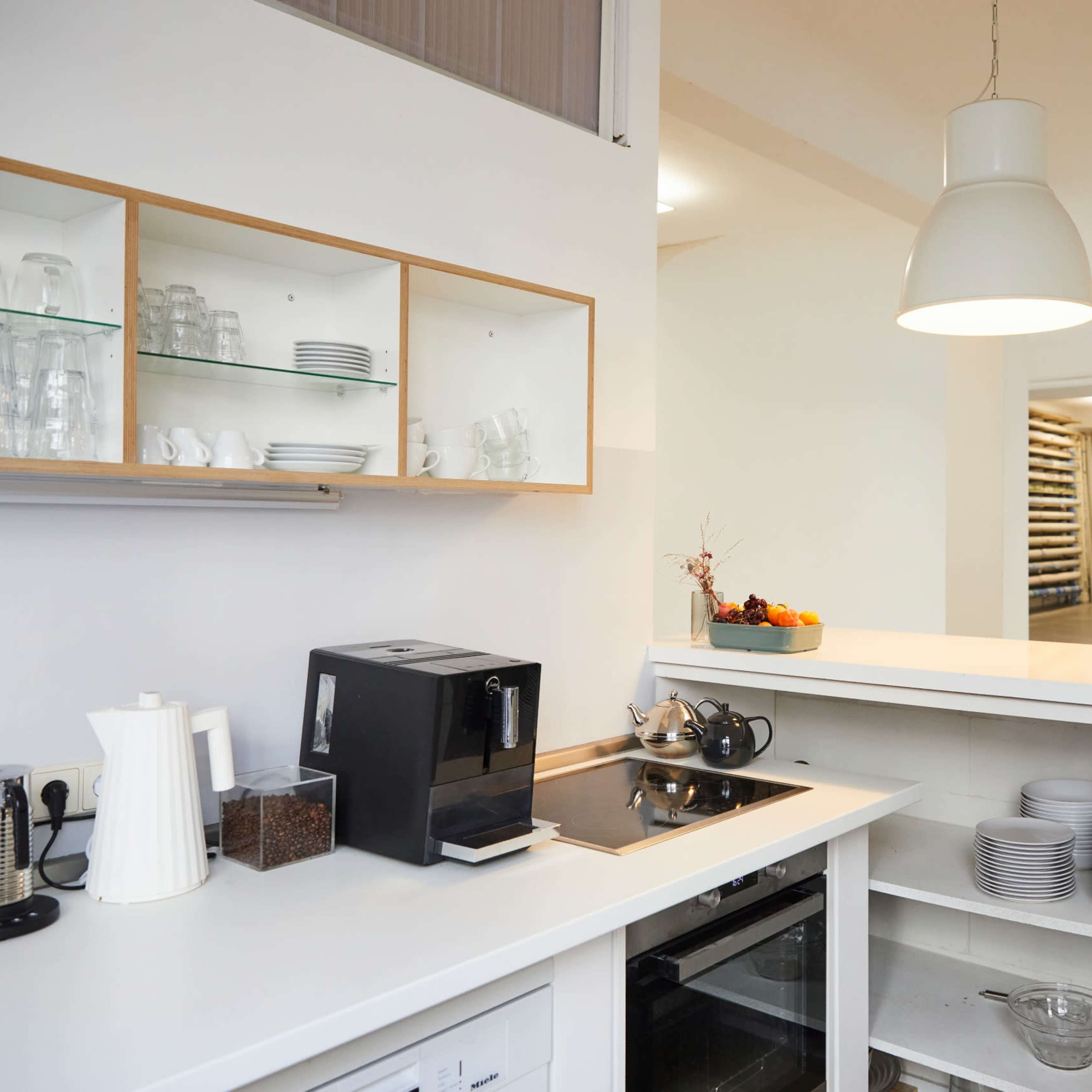 A modern kitchen area with a coffee maker, glassware on a shelf, and a neatly organized open space in the background.