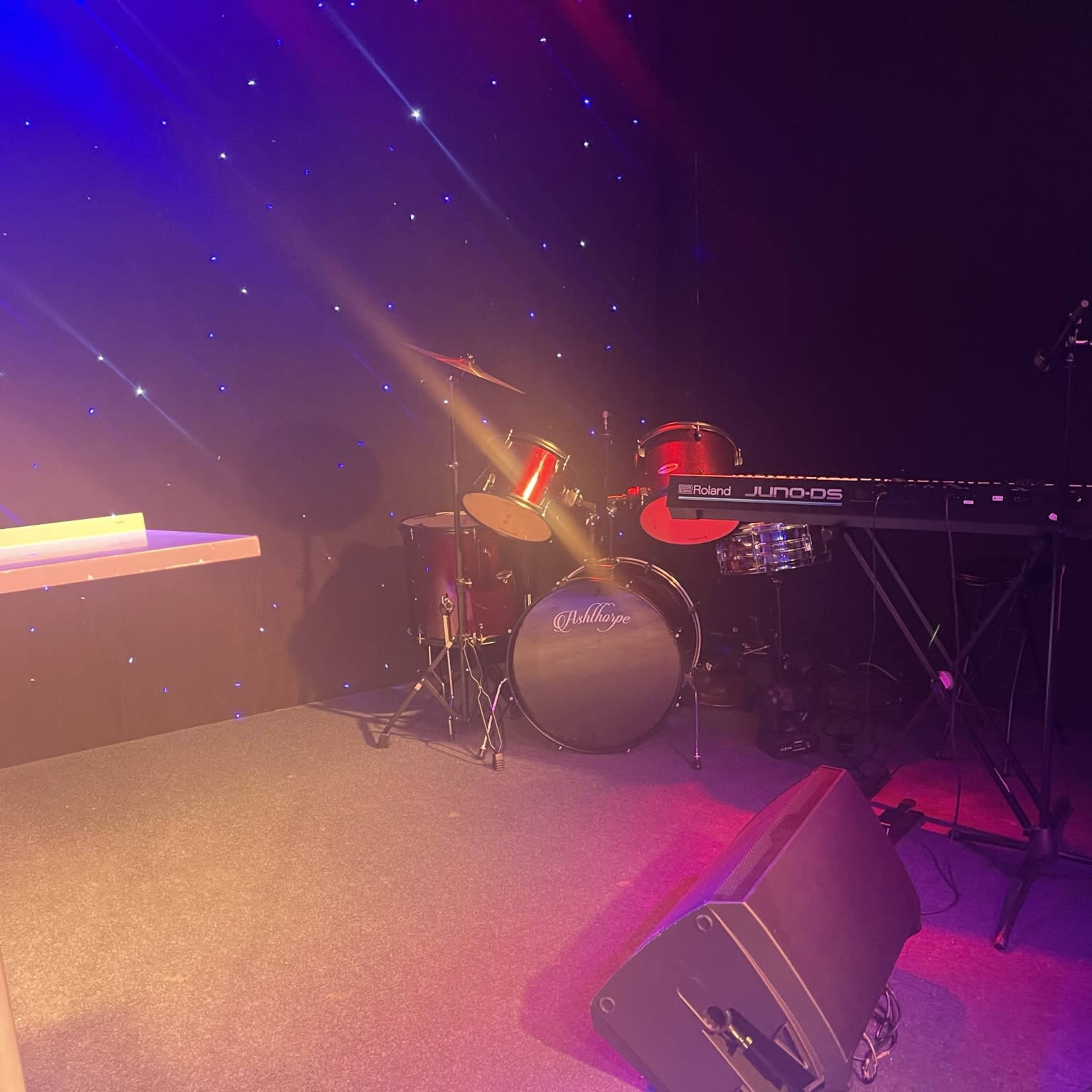 A drum set and a keyboard are positioned on a stage with a starry background.