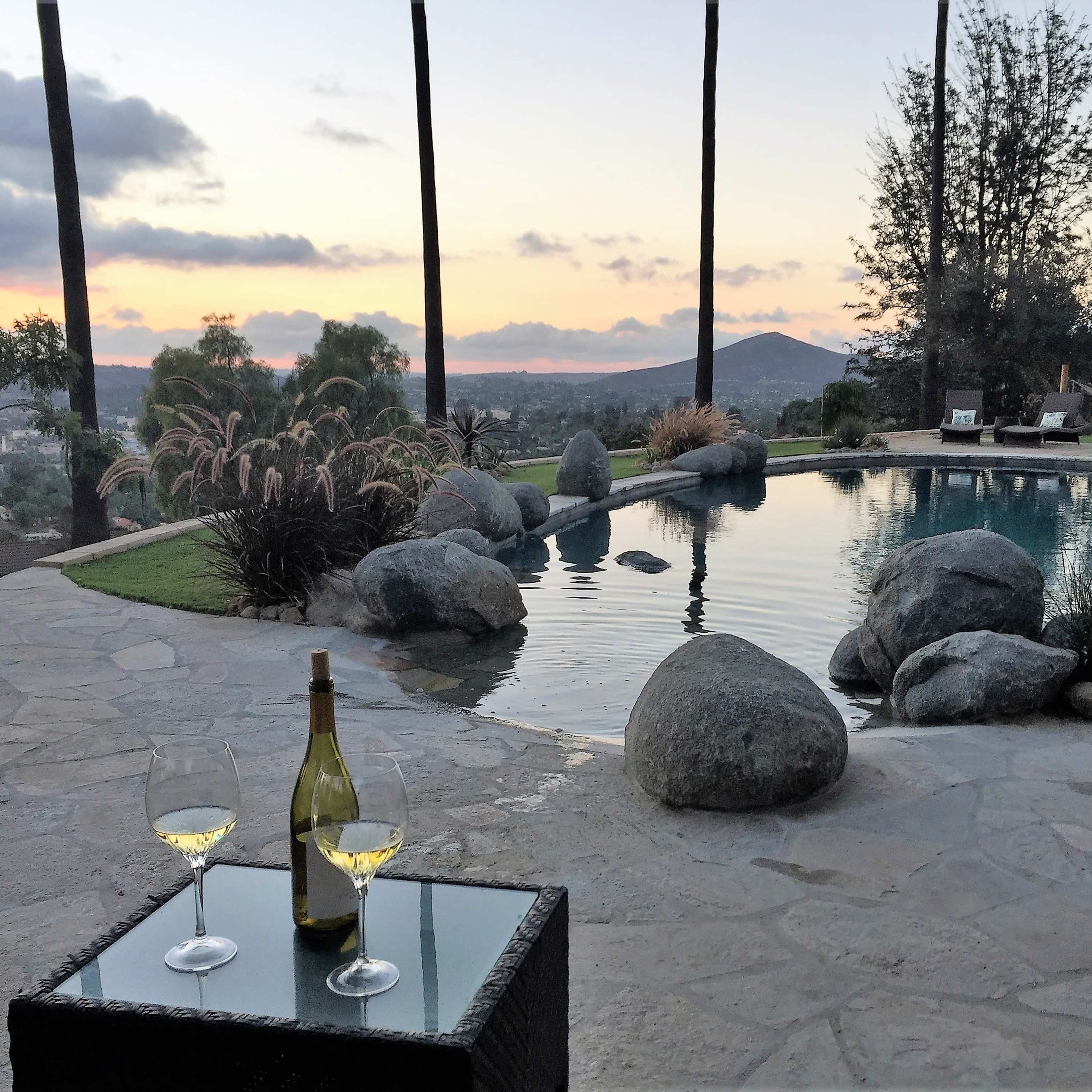 A pool area with large stones and a table holding two glasses of wine overlooks a sunset landscape with distant hills and palm trees.