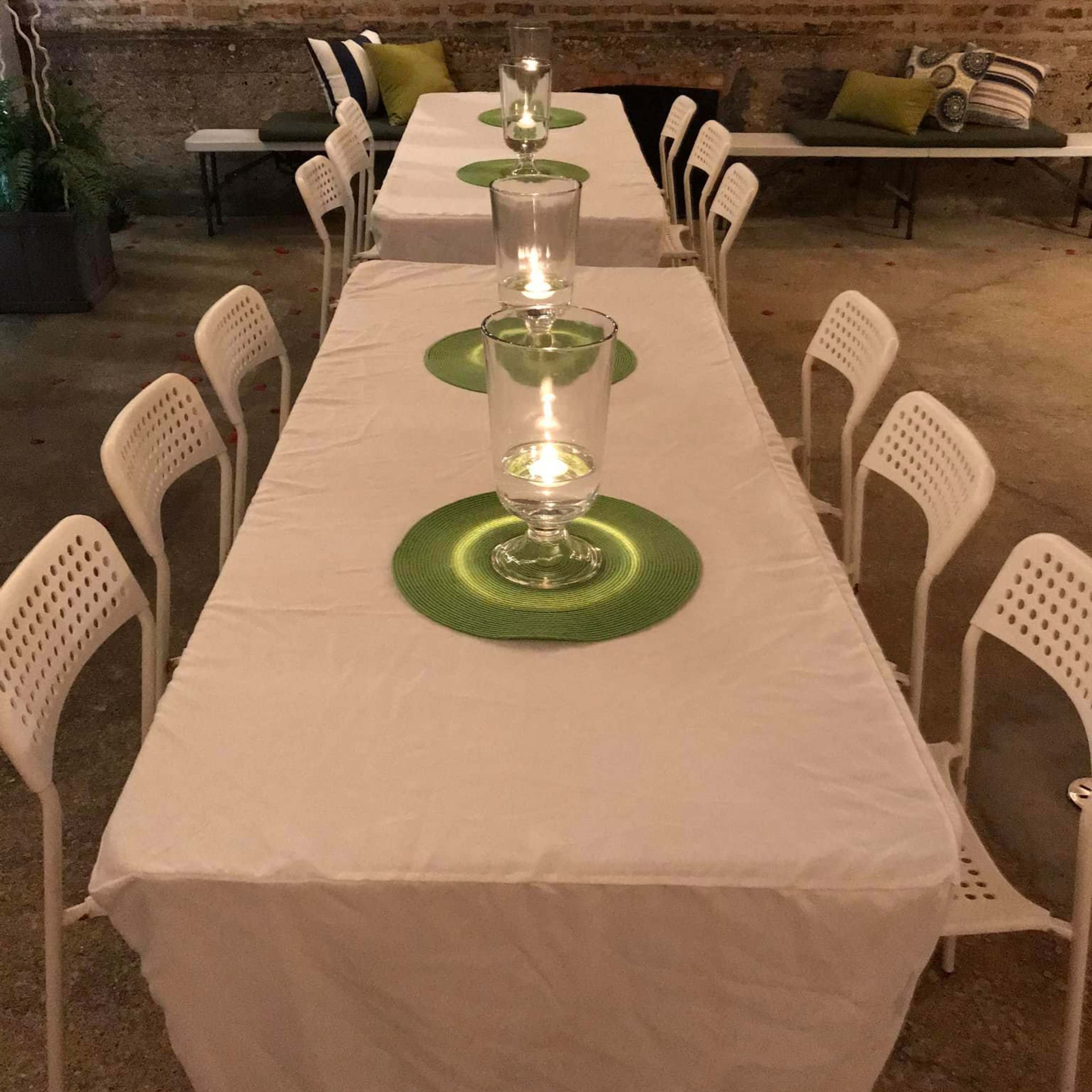Two long, white-covered tables set for a gathering, each adorned with green placemats and glass candle holders in the center, placed against a brick wall.