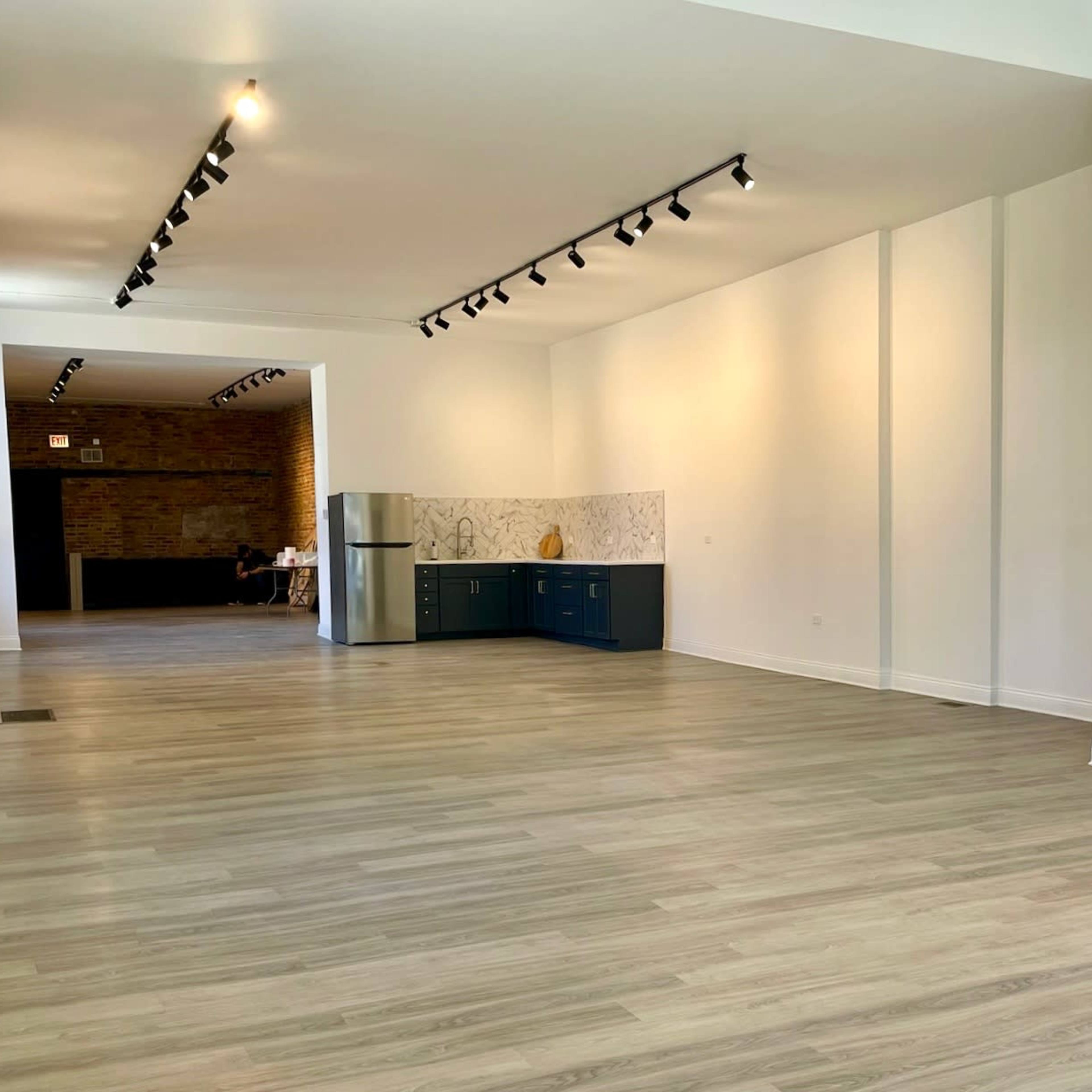 A spacious, empty room with light-colored walls and a wooden floor, featuring a kitchen area with blue cabinets at one end.