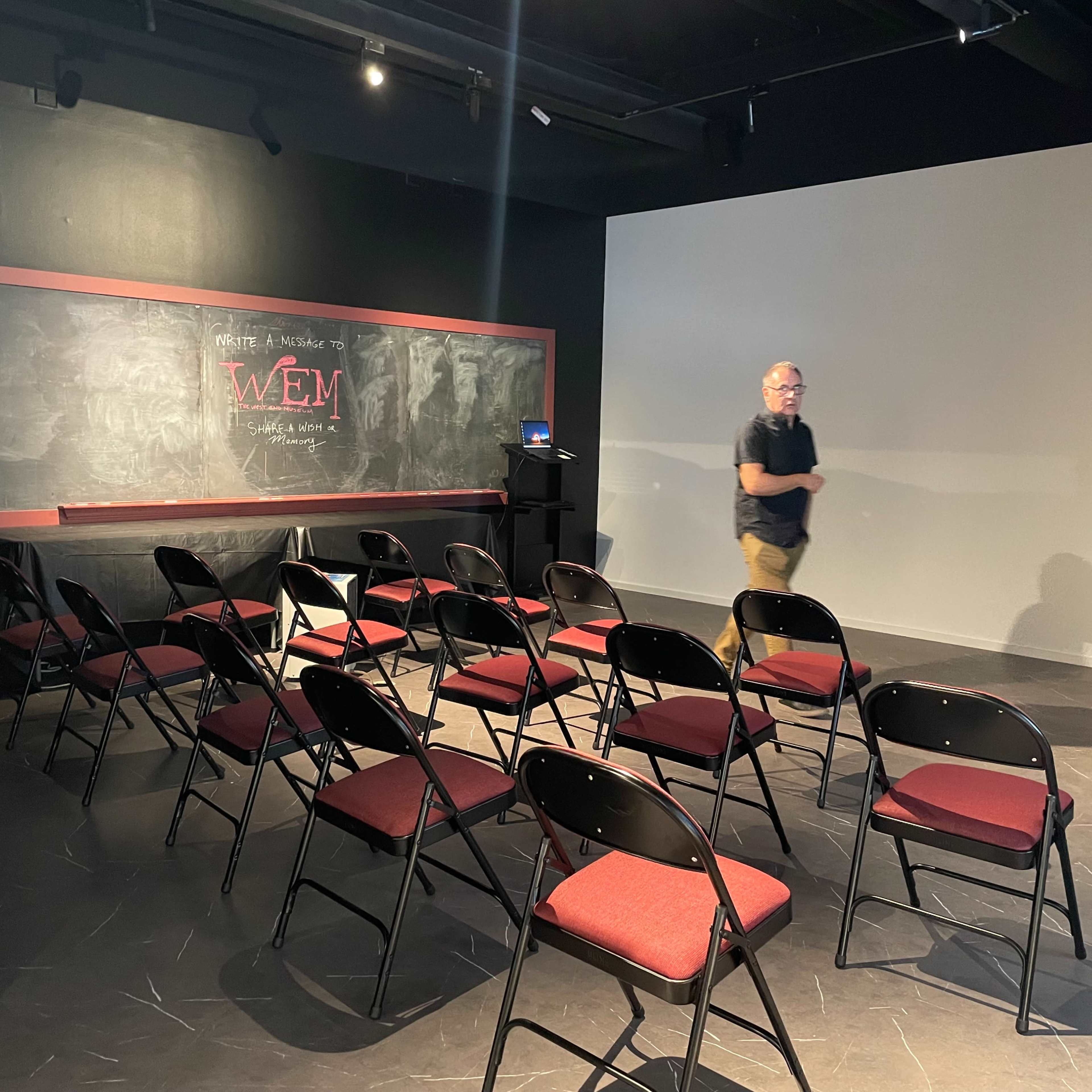 A man walks through a sparse, darkened room set up with rows of black chairs and a chalkboard at the front.