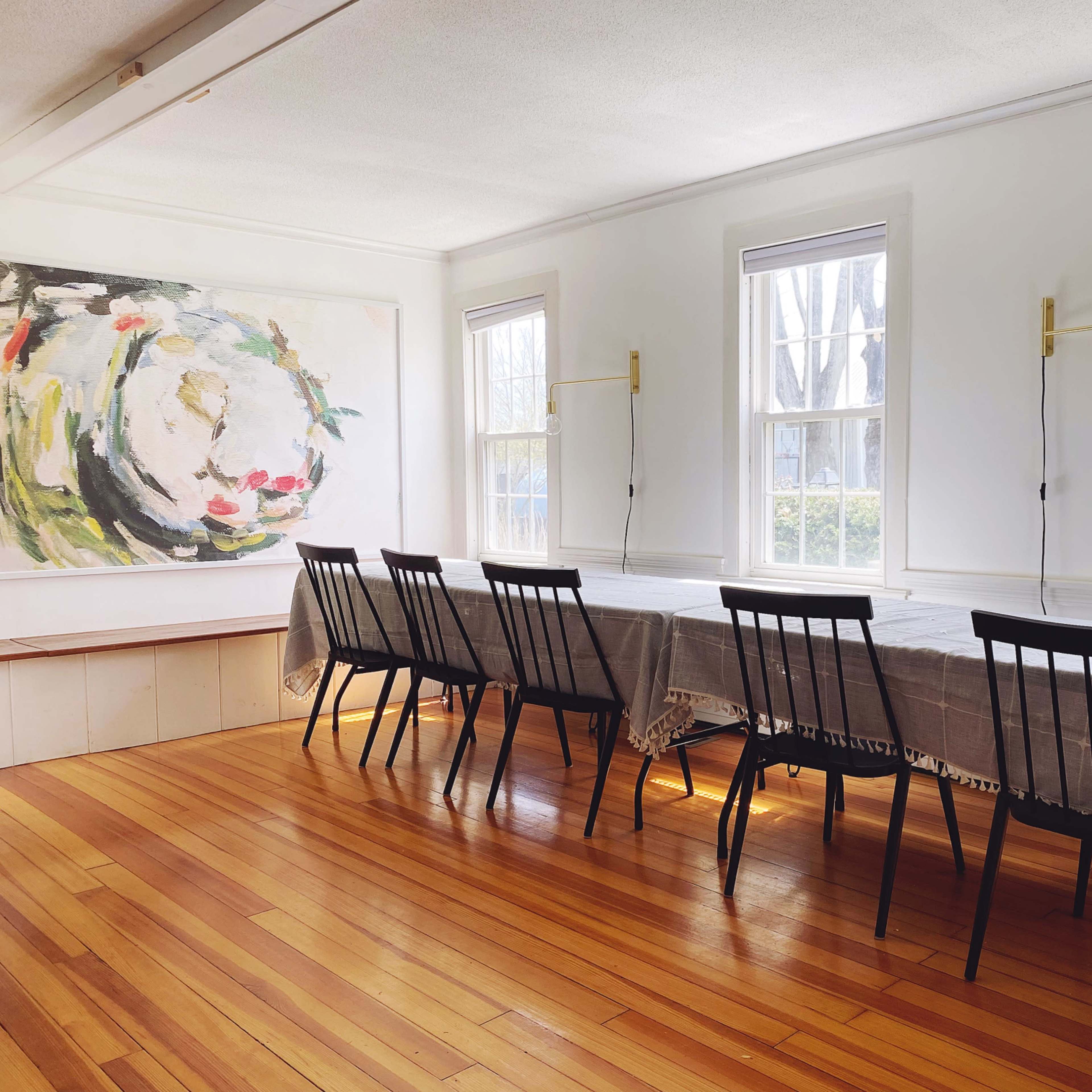 A long table with a gray tablecloth and eight black chairs is positioned in a well-lit dining room featuring large windows and a colorful painting on the wall.
