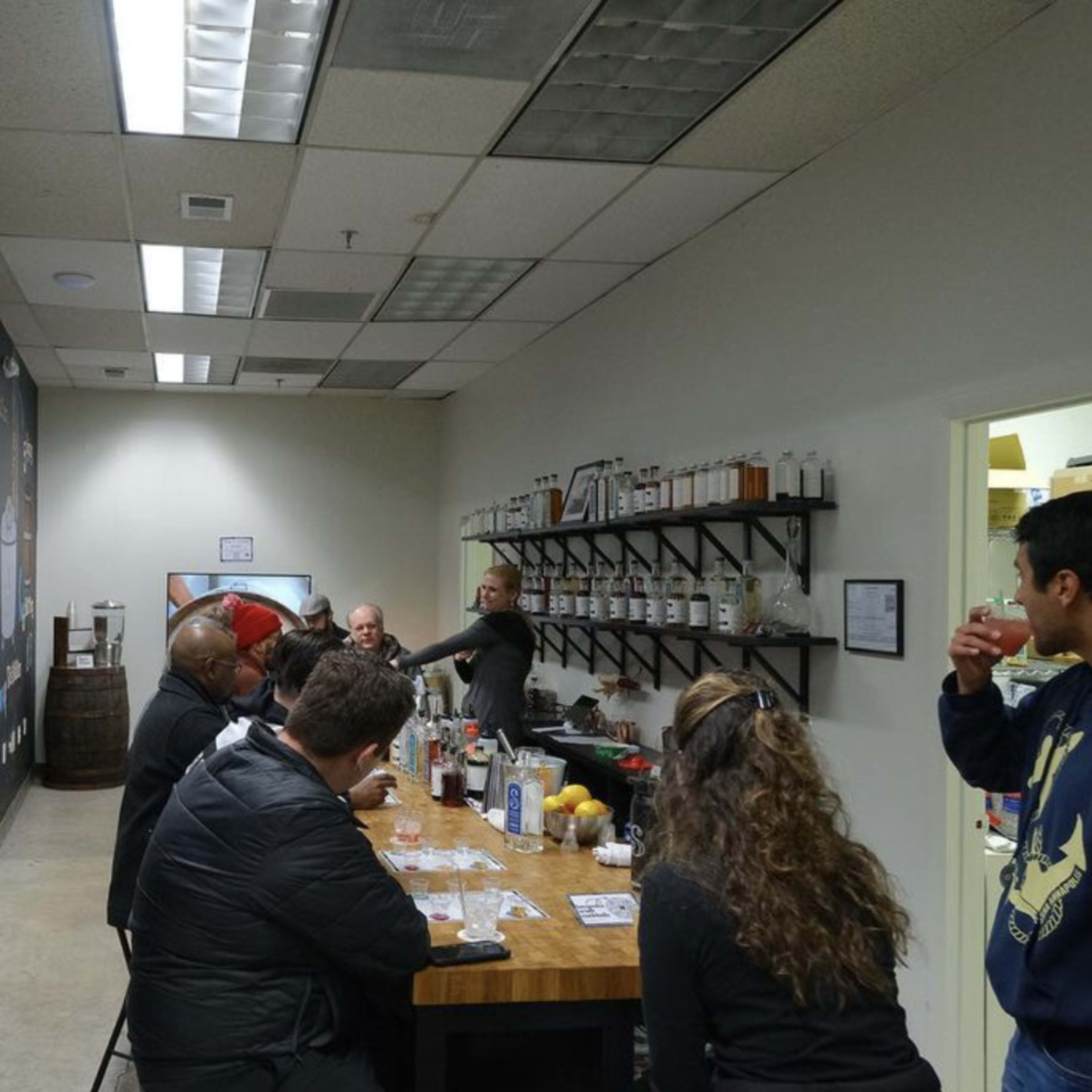 A group of people sits at a long wooden table in a brightly lit room with shelves of bottles and a black chalkboard wall.