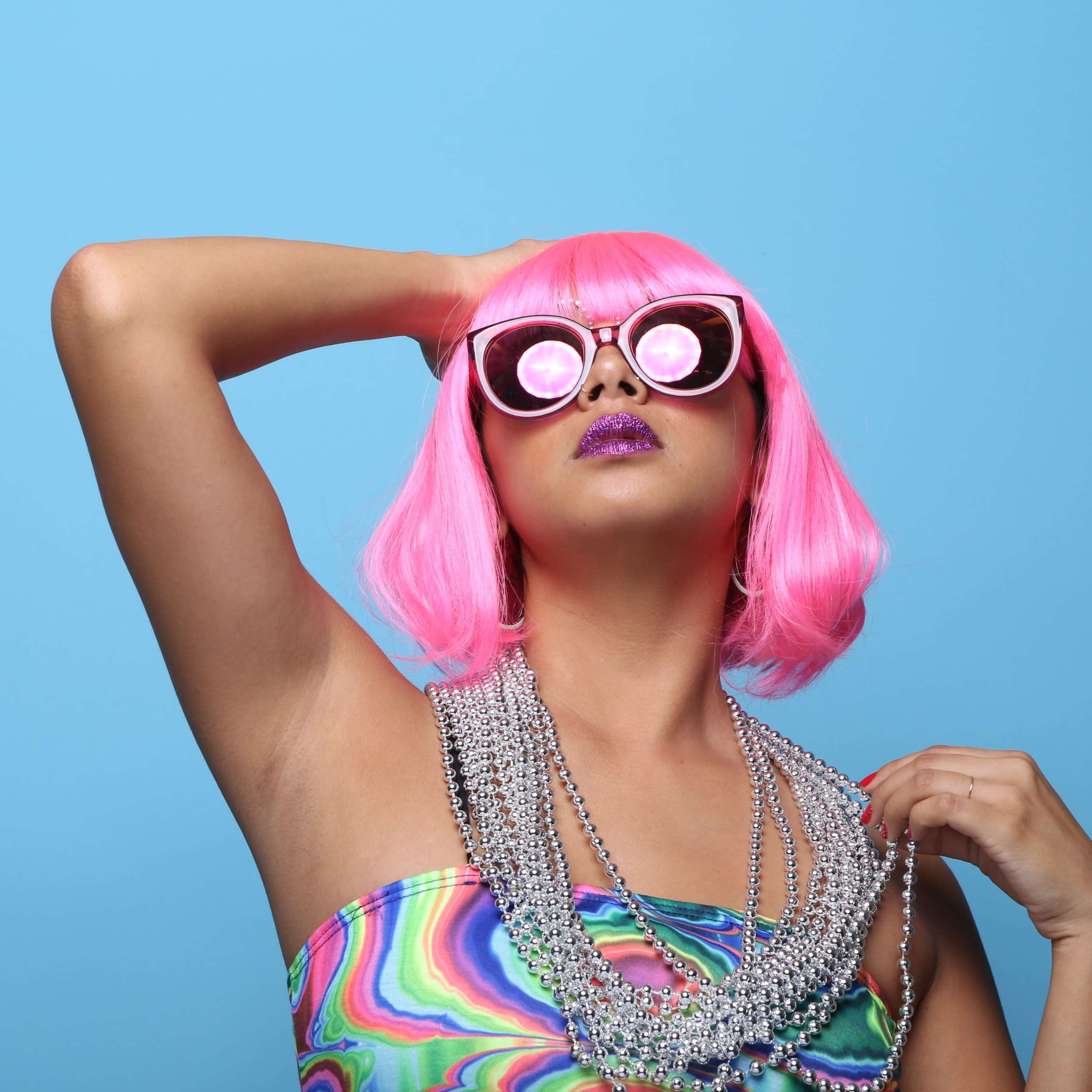 A person with pink hair and oversized sunglasses poses confidently against a blue background while wearing a colorful outfit and holding a necklace.