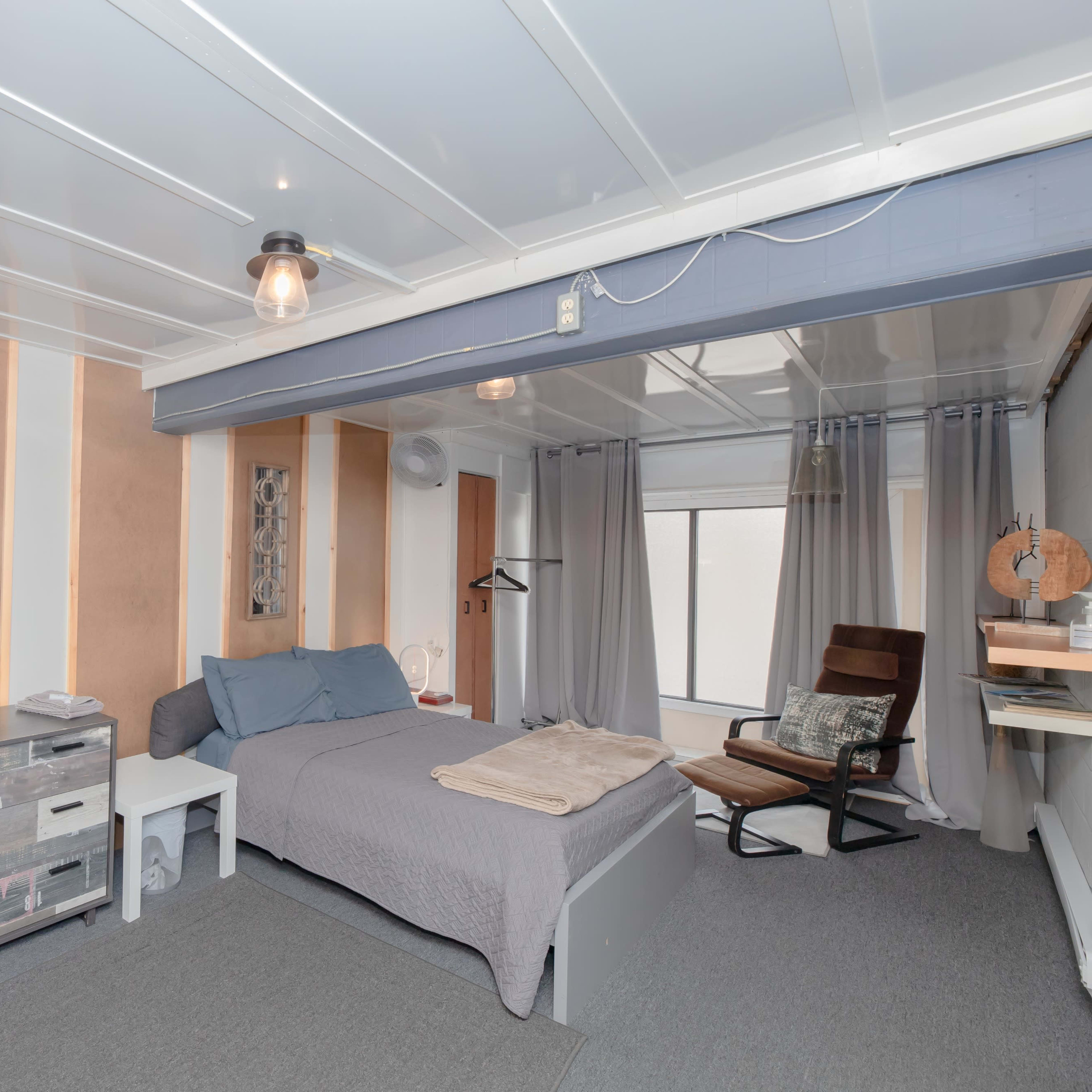 A contemporary bedroom with a bed, a wooden dresser, a modern chair, and large windows covered by sheer curtains.