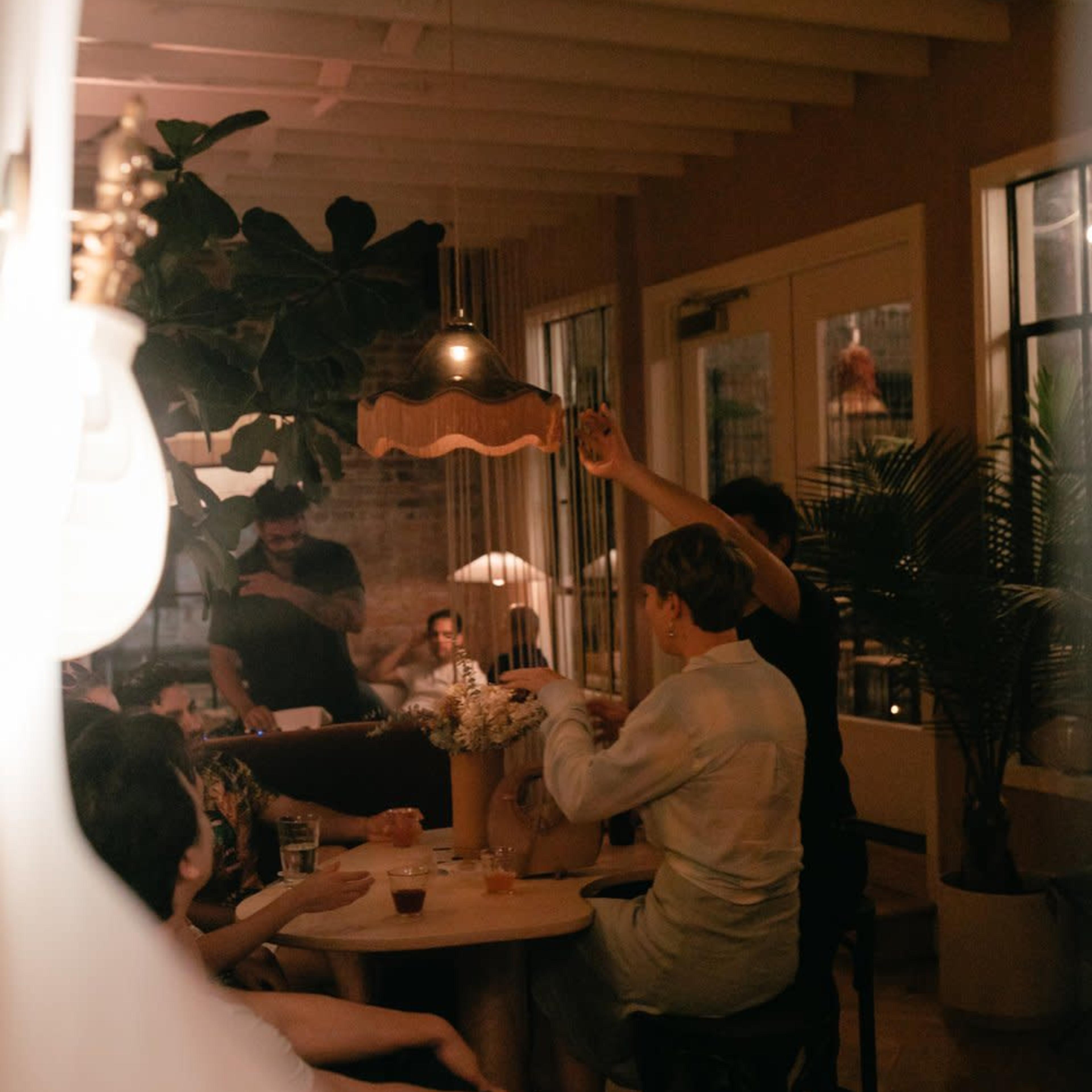 A group of people sits around a table in a warm, dimly lit interior, sharing drinks and conversation.