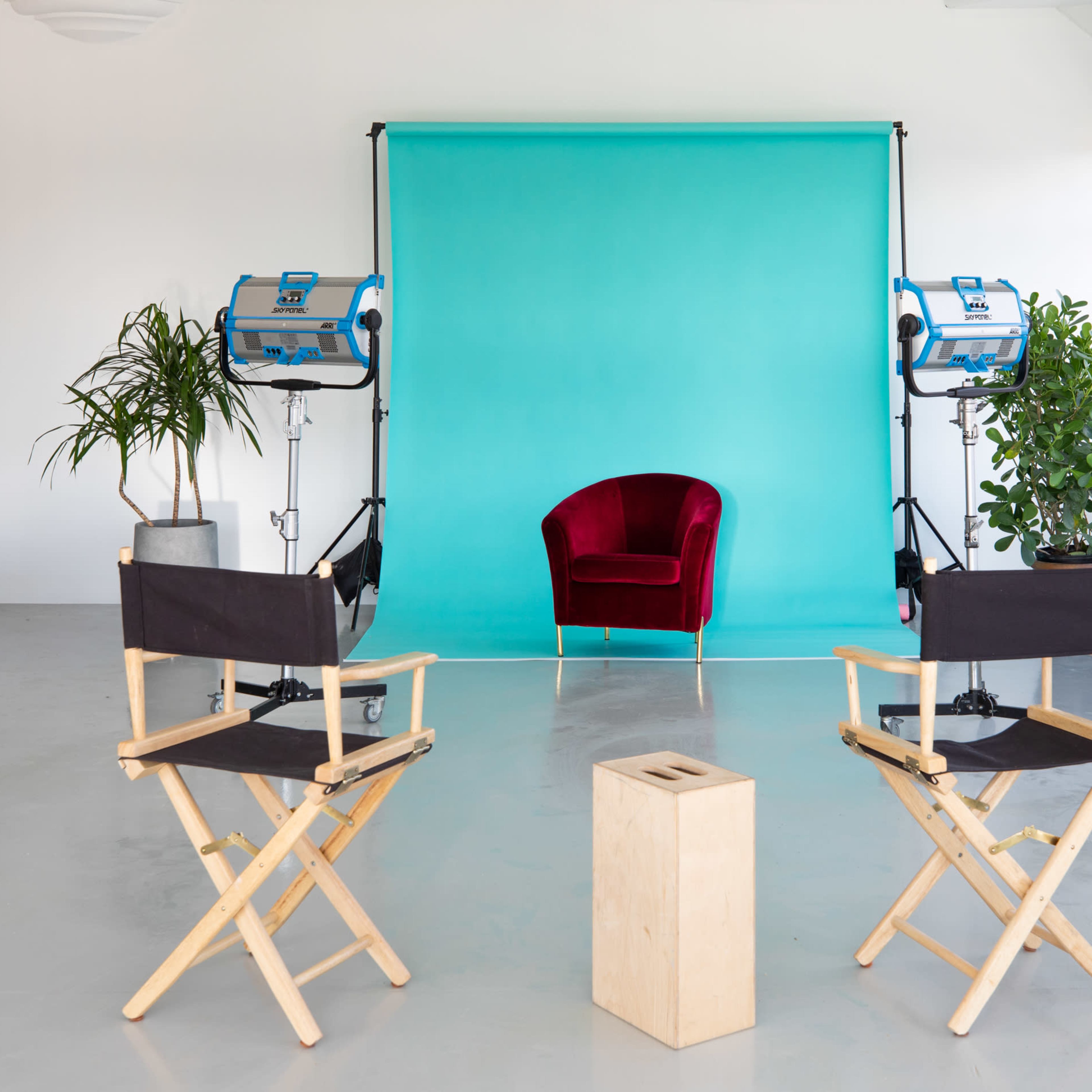 A photo studio featuring a turquoise backdrop, a red velvet chair, two studio lights, and two director's chairs positioned around a wooden stool.