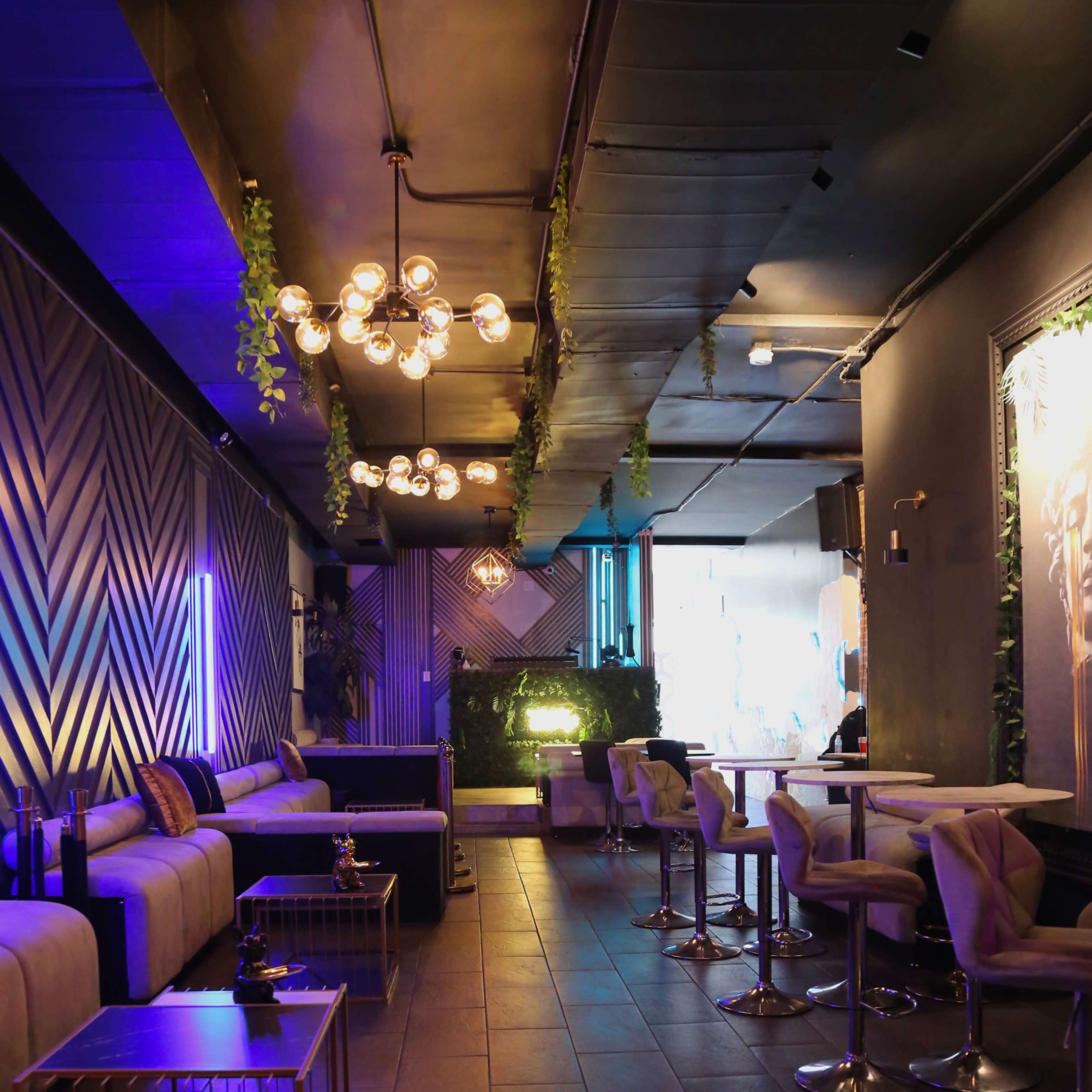 A dimly lit lounge with modern seating, decorative lighting, and plants, leading to an illuminated area at the back.