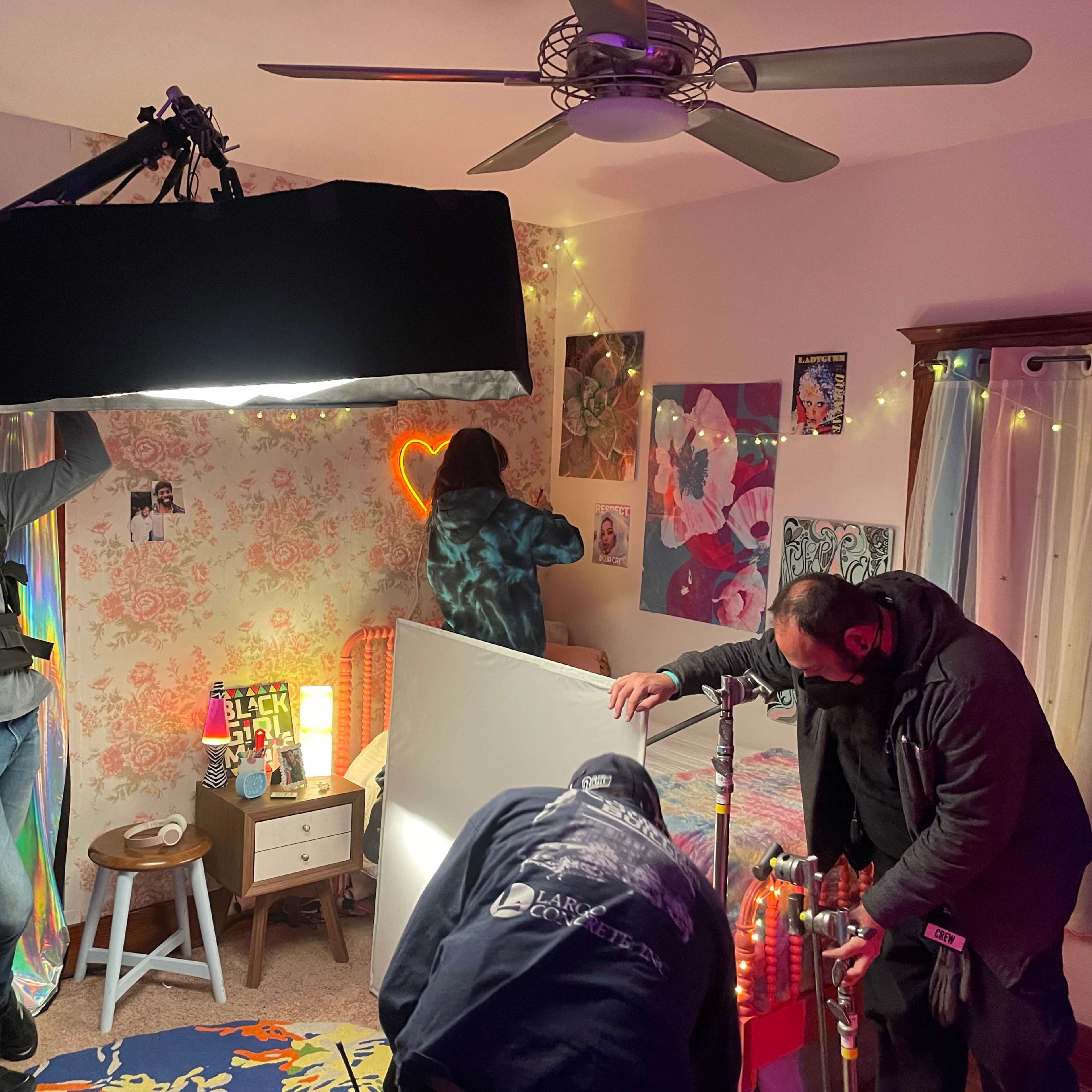 A film crew sets up equipment in a brightly decorated bedroom with posters and string lights.