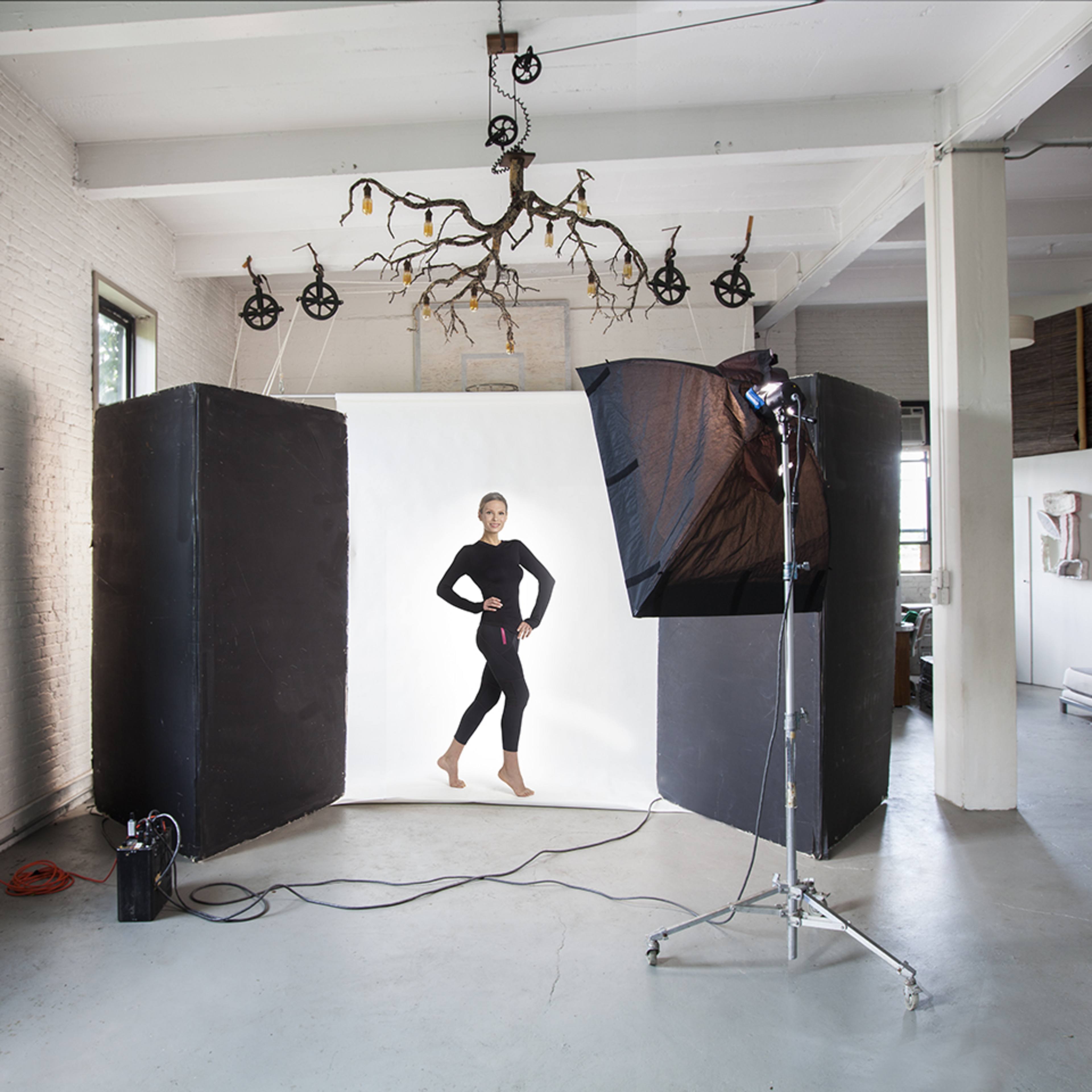 A model poses in a photography studio with a backdrop while studio lights and equipment are set up around her.
