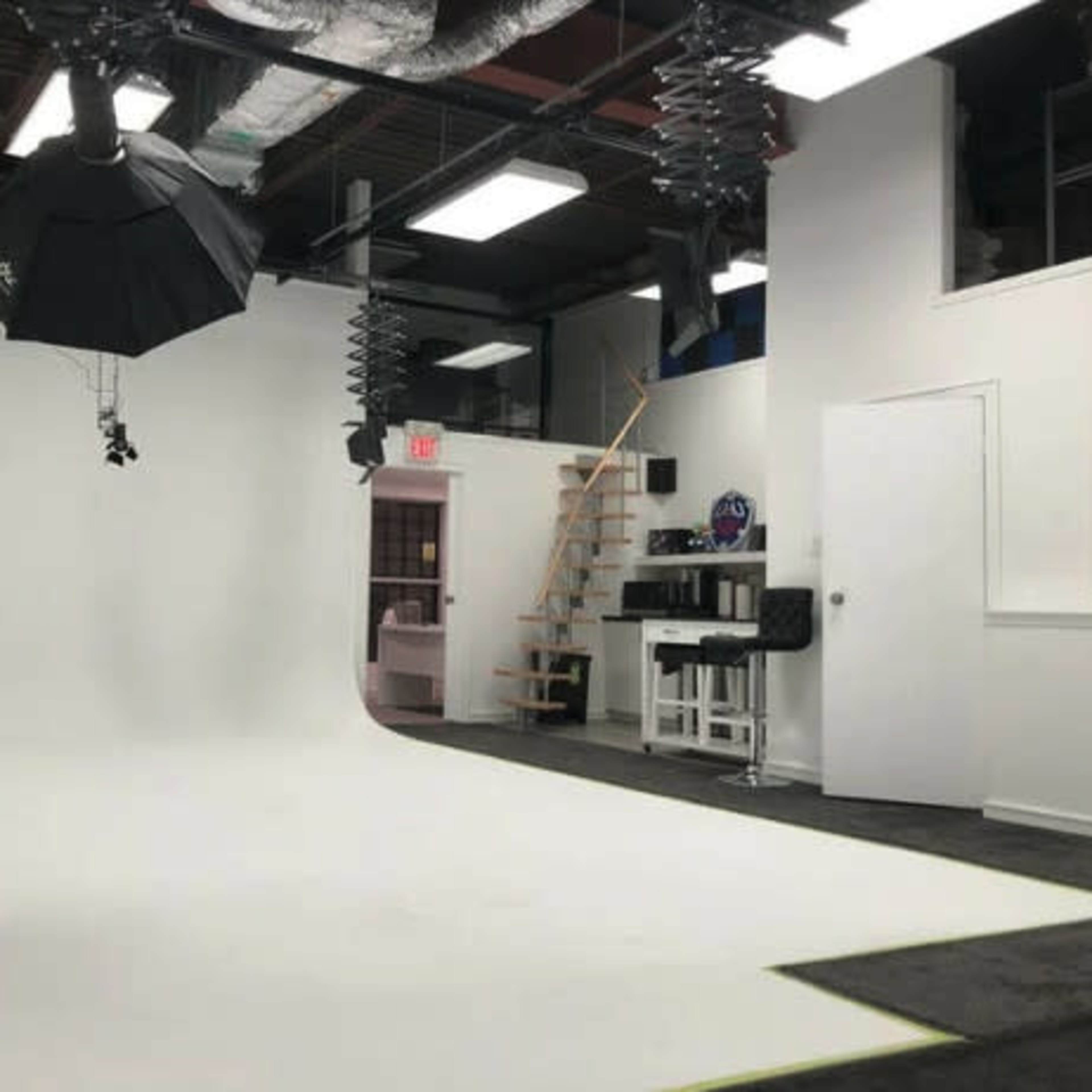 A photography studio with a curved white backdrop, overhead lighting, and a staircase leading to an upper level.