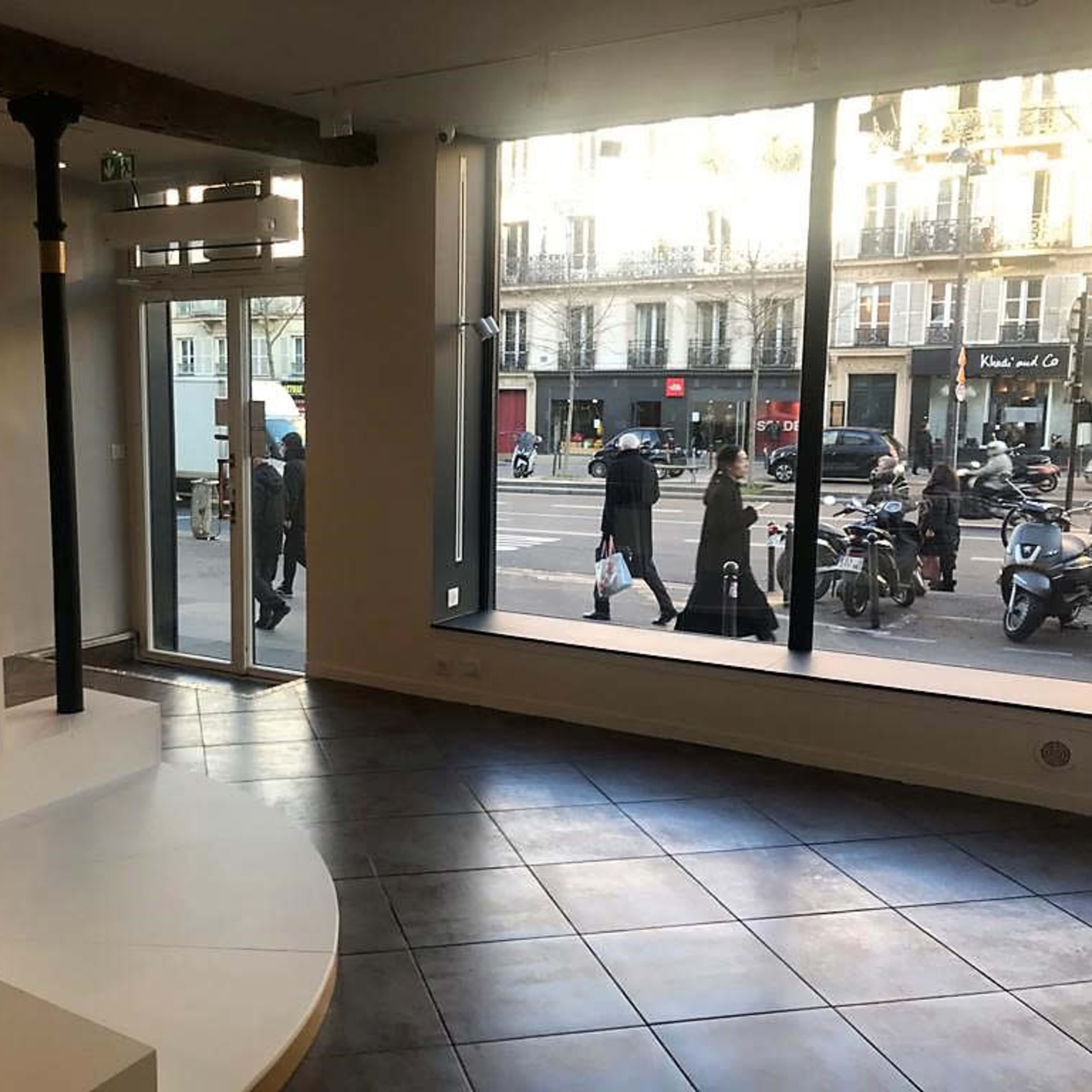 A modern, minimalist interior with a circular white platform and large windows overlooking a street with people and parked motorcycles outside.