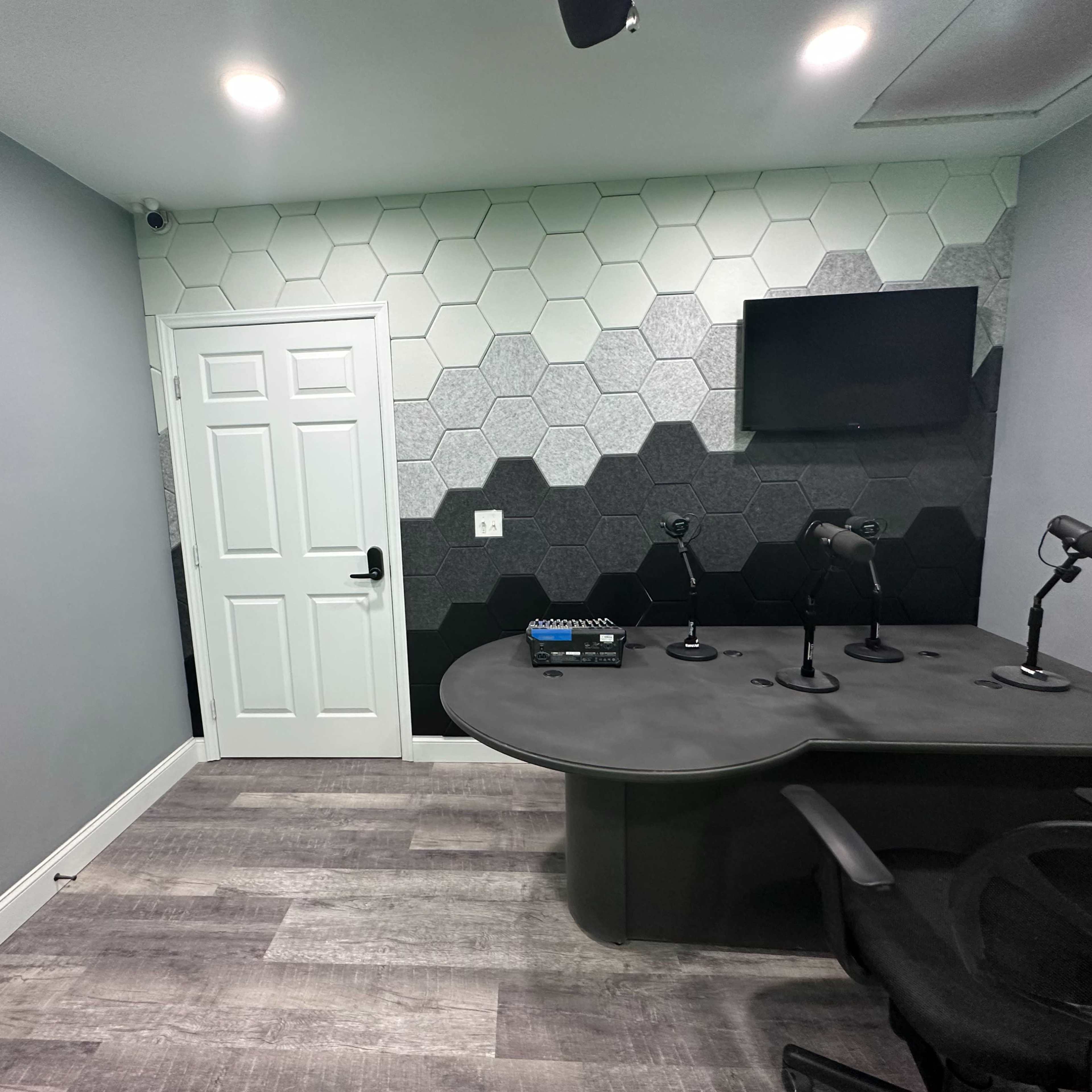 A modern recording studio with a hexagonal-patterned accent wall, a desk with multiple microphone stands, and a wall-mounted television.