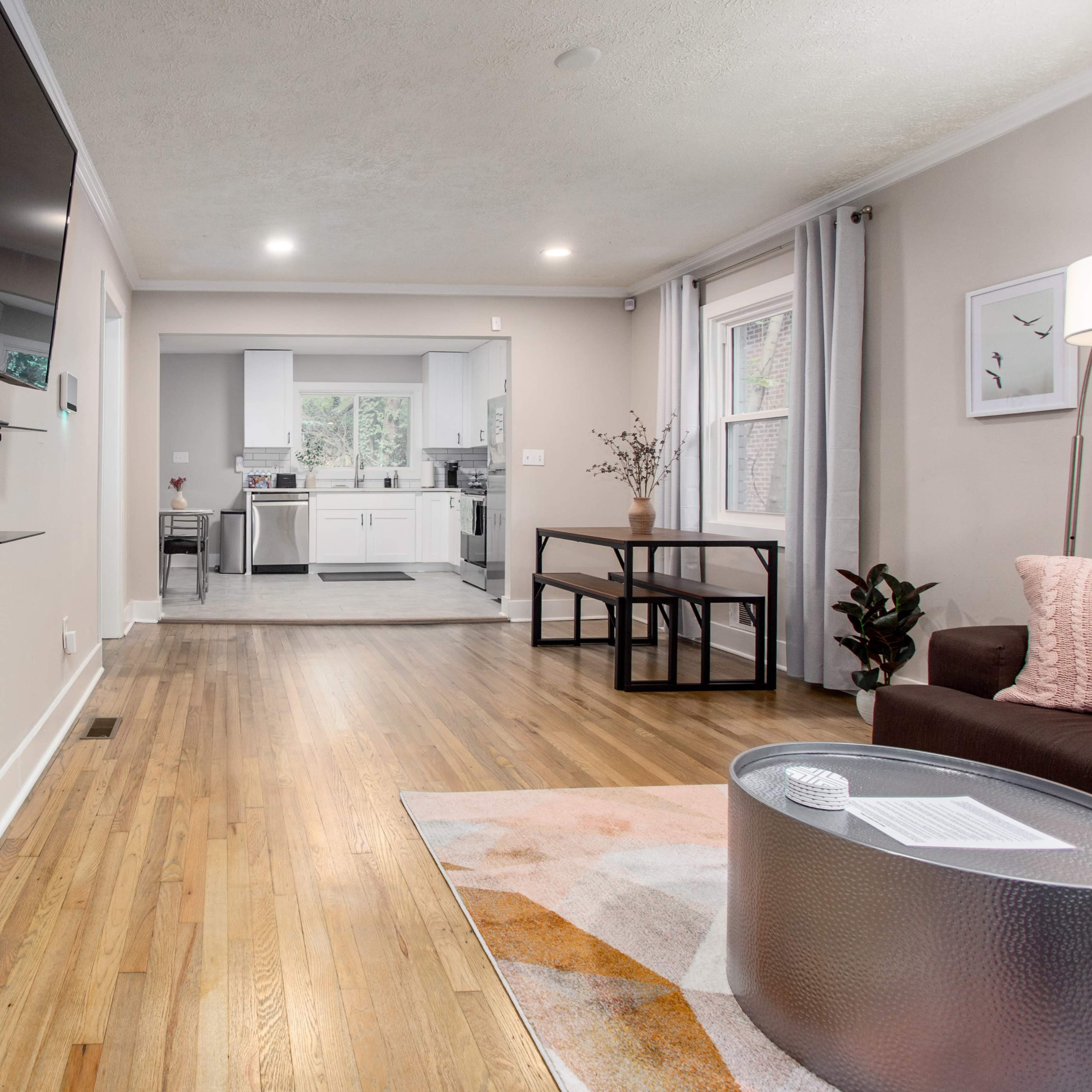 A living room with a wall-mounted television and a view into a kitchen area, featuring hardwood floors and a small dining table.
