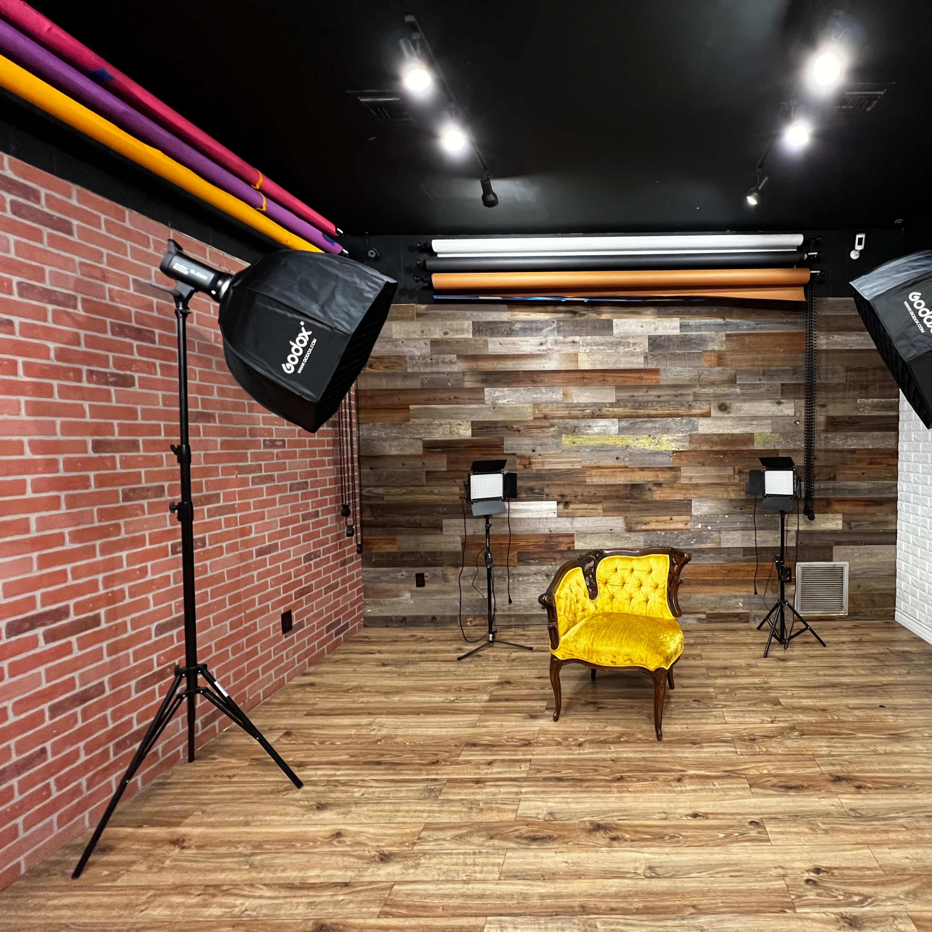 A photography studio featuring a yellow upholstered chair positioned in the center, flanked by two softbox lighting setups against a backdrop of textured wooden panels and white brick walls.