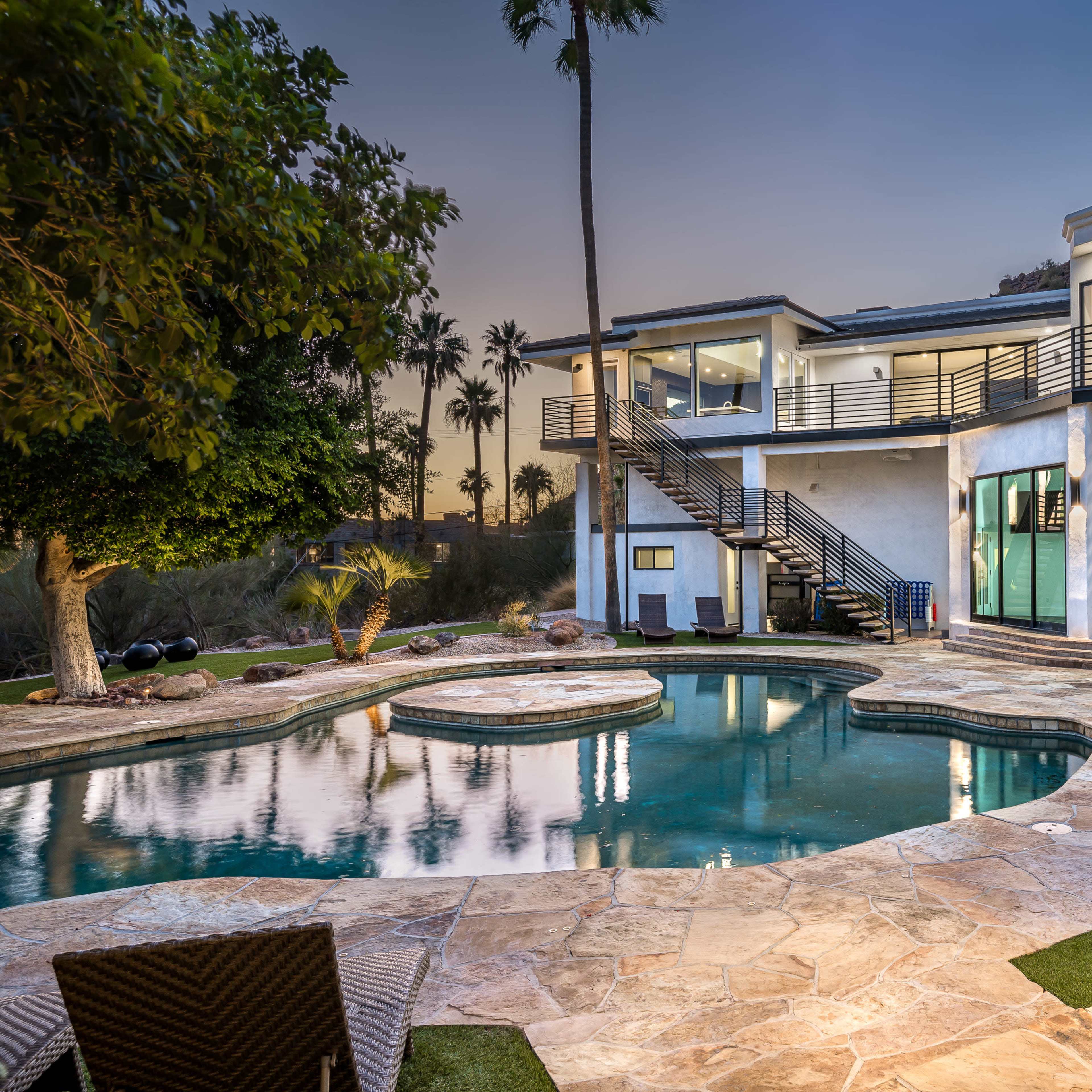 A modern house with large glass windows overlooks a swimming pool surrounded by stone patio and lounge chairs, set against a backdrop of palm trees and a twilight sky.