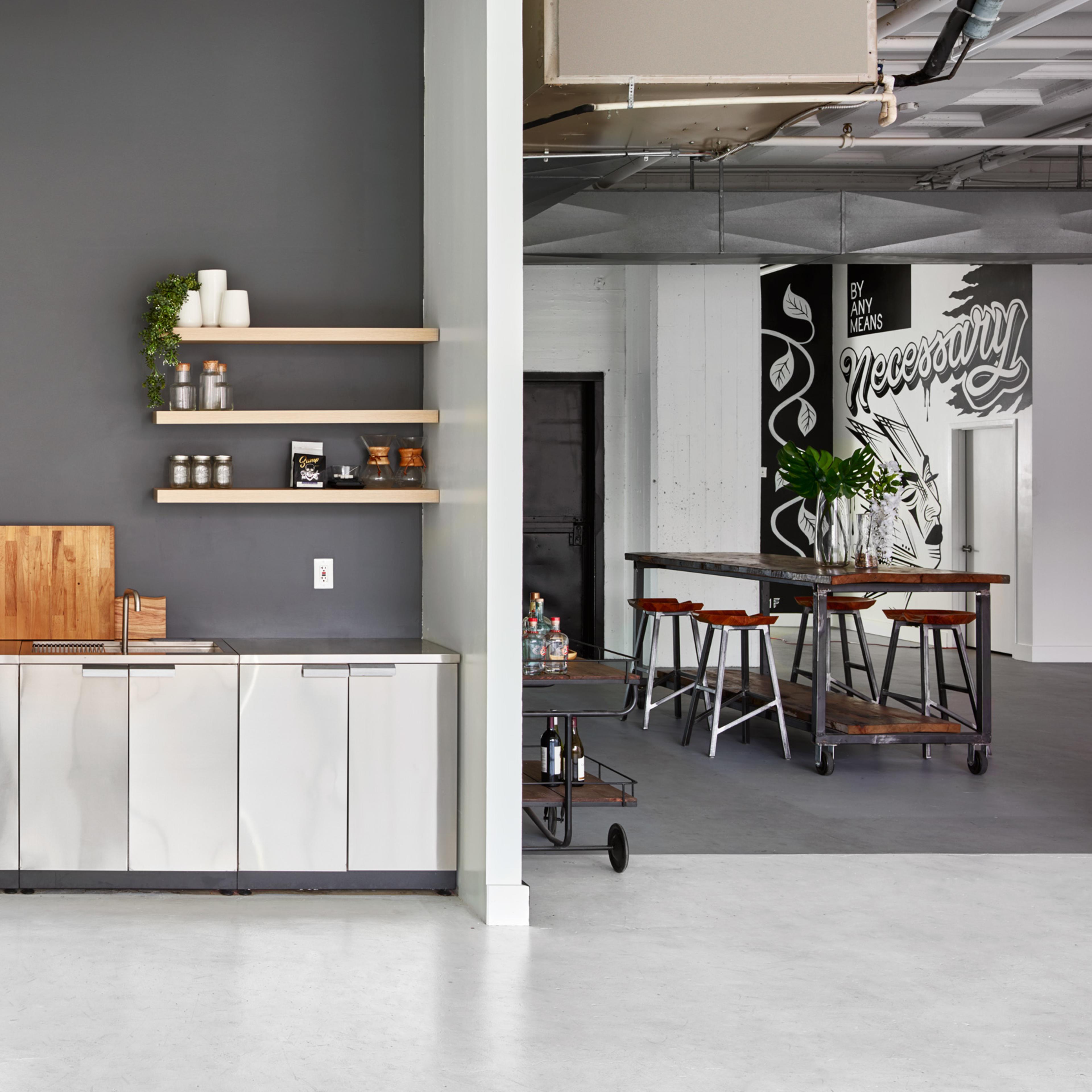 A modern kitchen area featuring stainless steel cabinets, a refrigerator, a microwave, and wooden shelving against a gray wall.