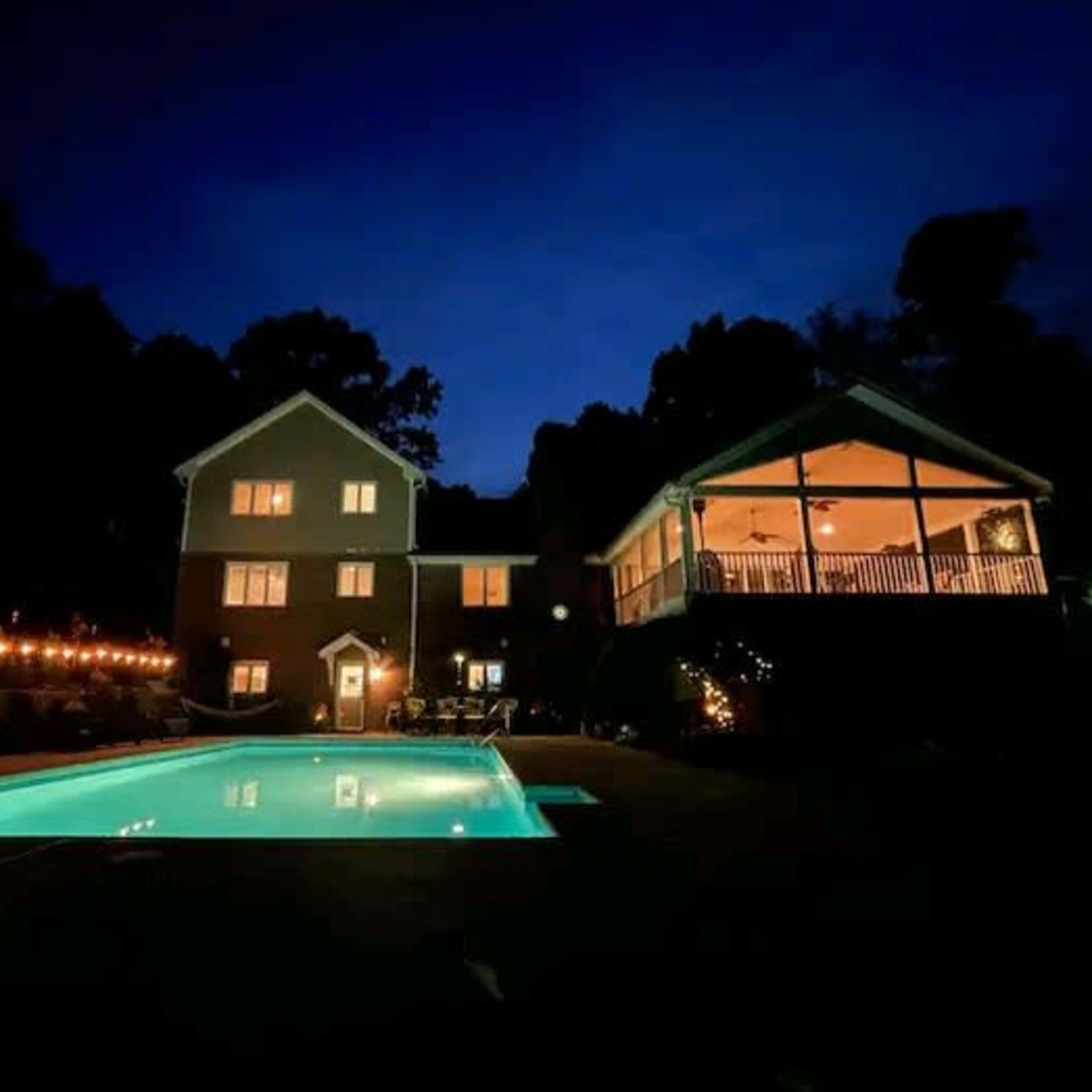 A well-lit pool area is surrounded by a two-story house and a covered porch at dusk.
