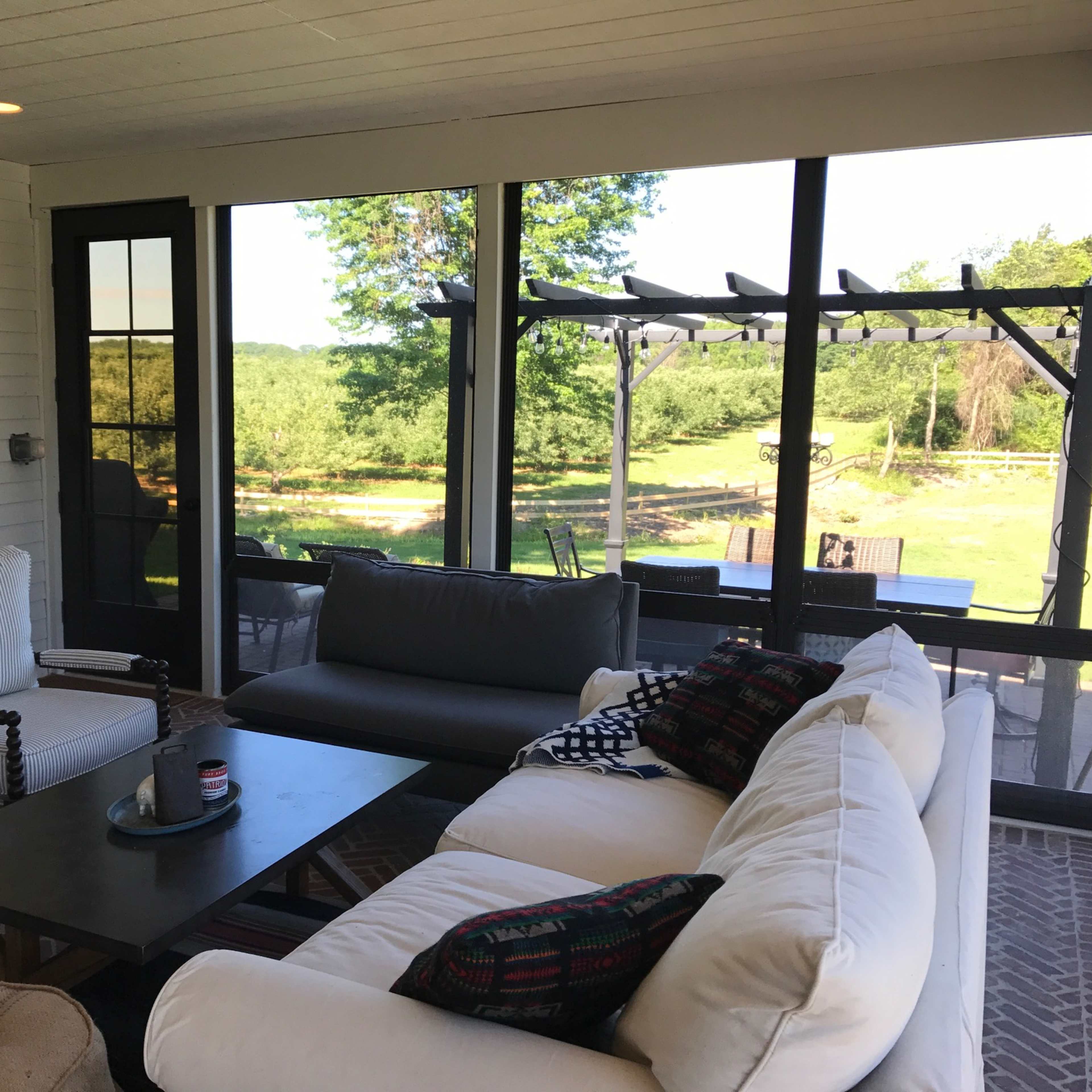 A screened-in porch features white and dark furniture, a coffee table, and large windows overlooking a green landscape.
